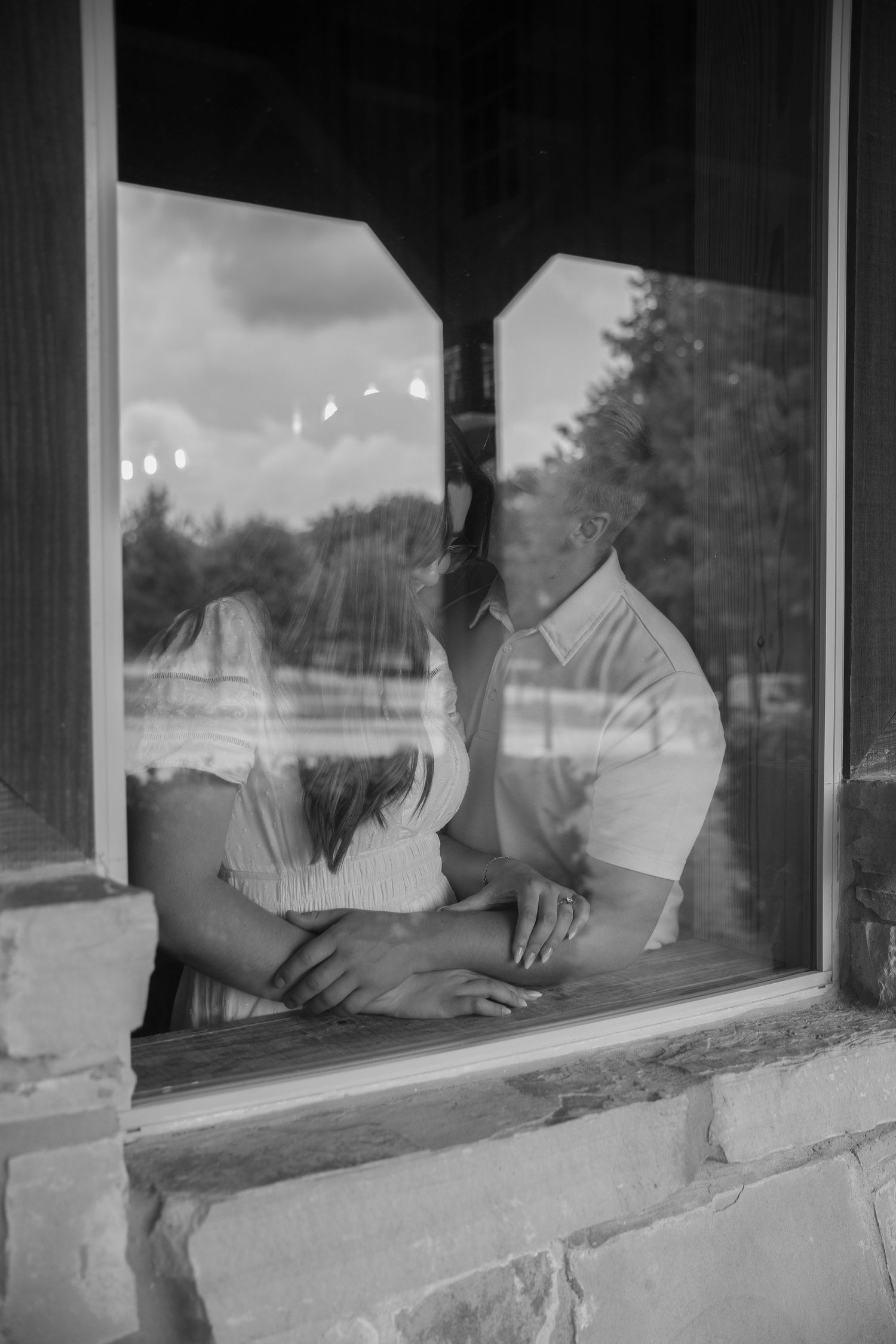 Black and white photo of a couple holding hands while sitting together beside a rustic window.