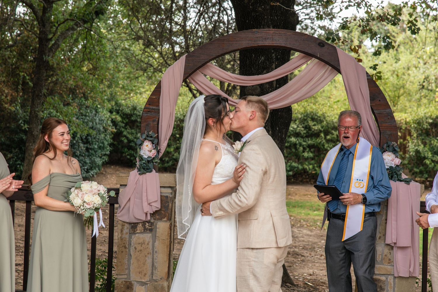 Bride and groom share a kiss beneath a rustic wooden wedding arch draped in blush fabric as a bridesmaid and officiant look on.