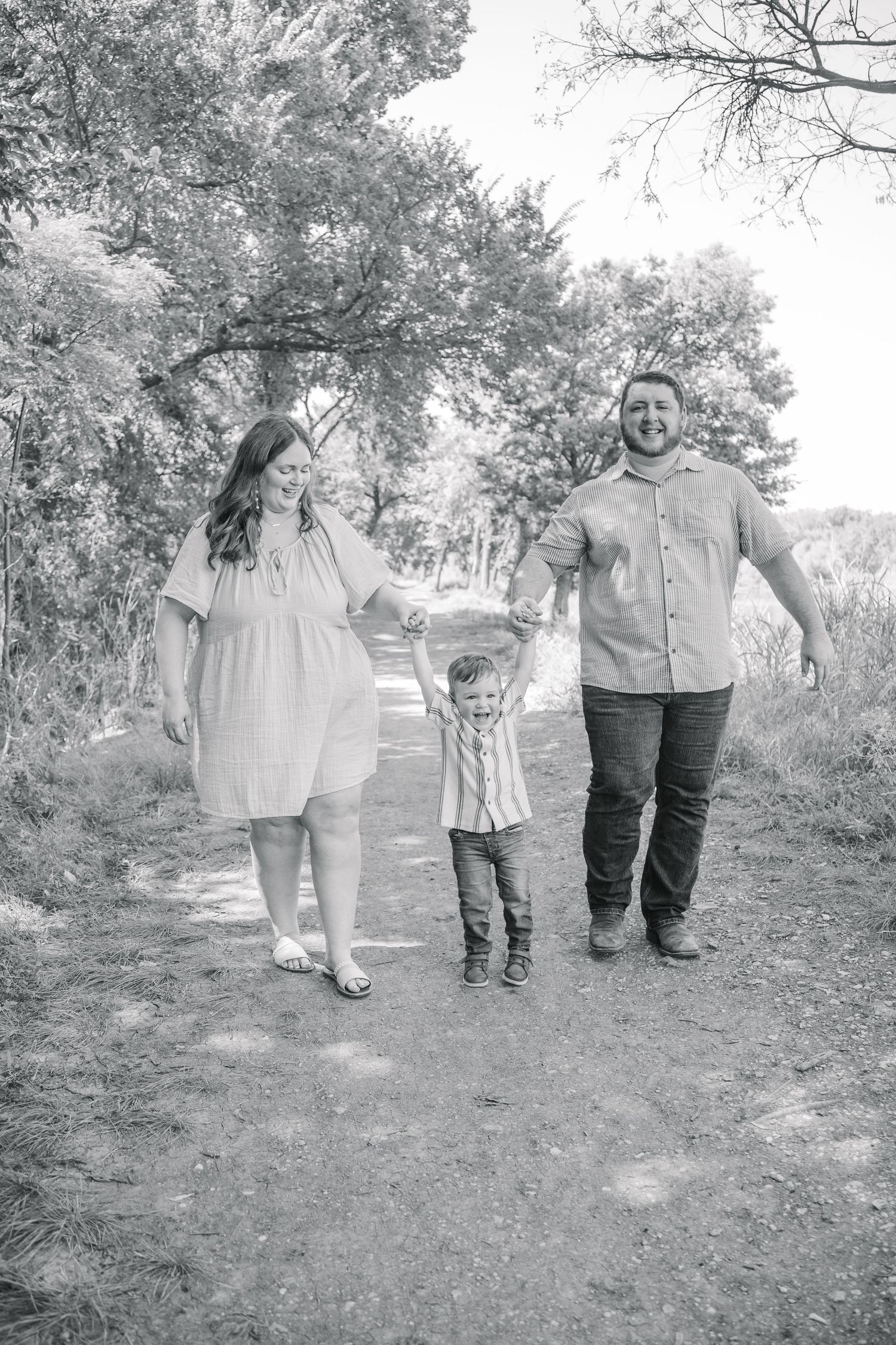 Black and white photo of a happy family with a young child holding parents hands walking on a scenic outdoor path.
