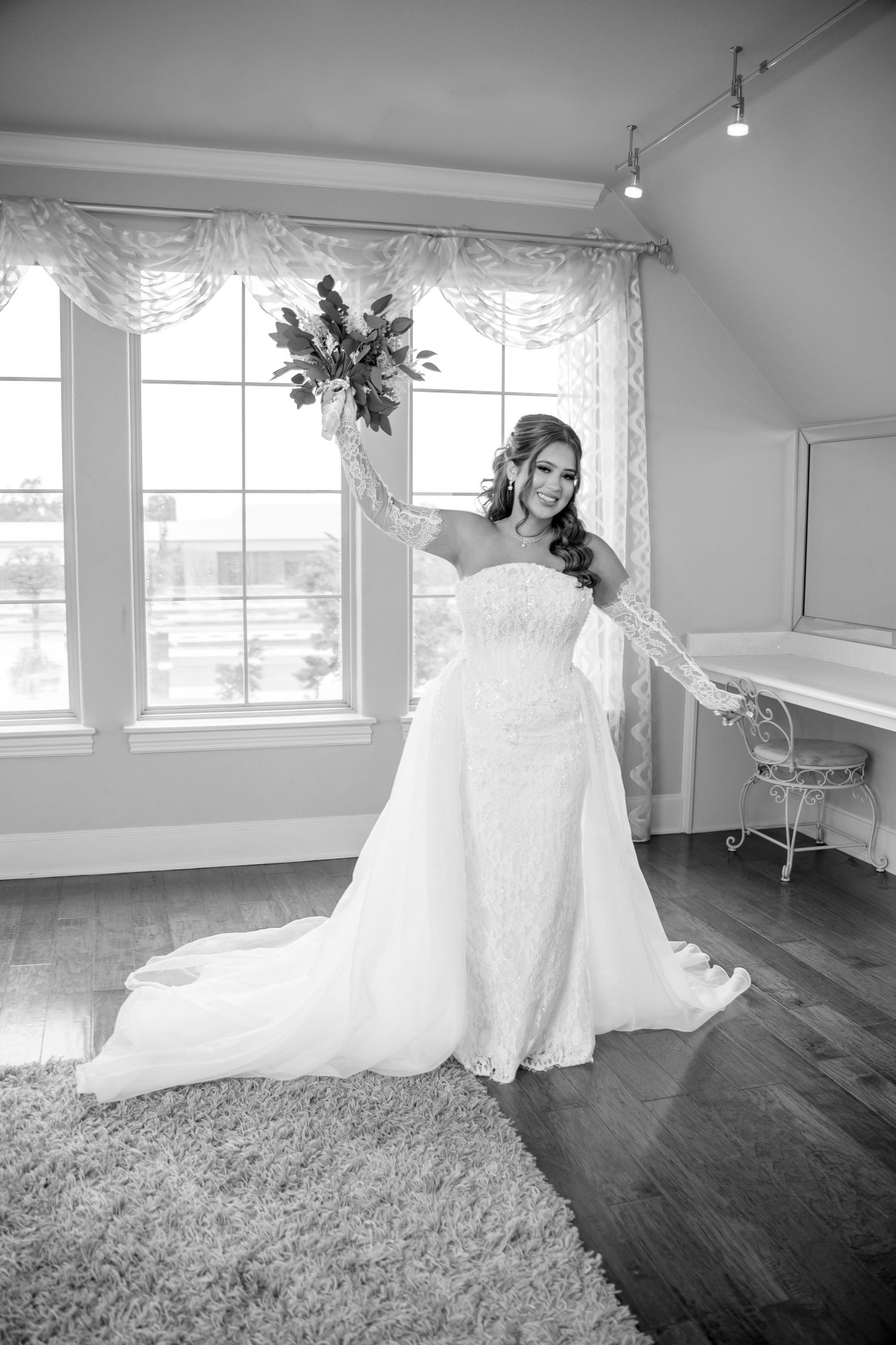 Bride in strapless lace gown holds bouquet aloft in bright bridal suite, black and white photo.