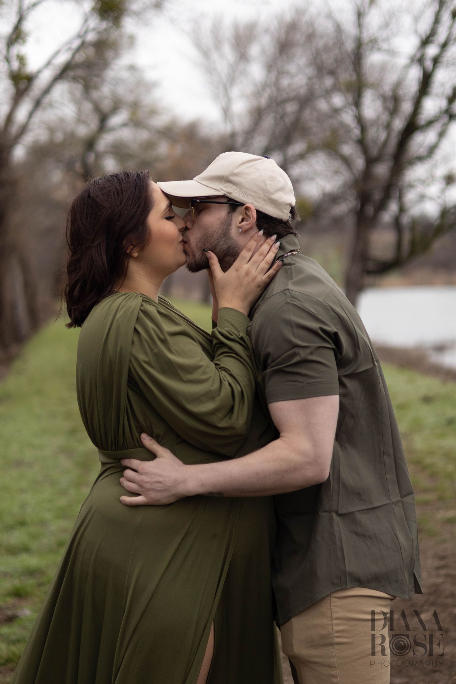 Pregnant woman in olive green dress shares a kiss with partner in beige cap during outdoor maternity shoot.