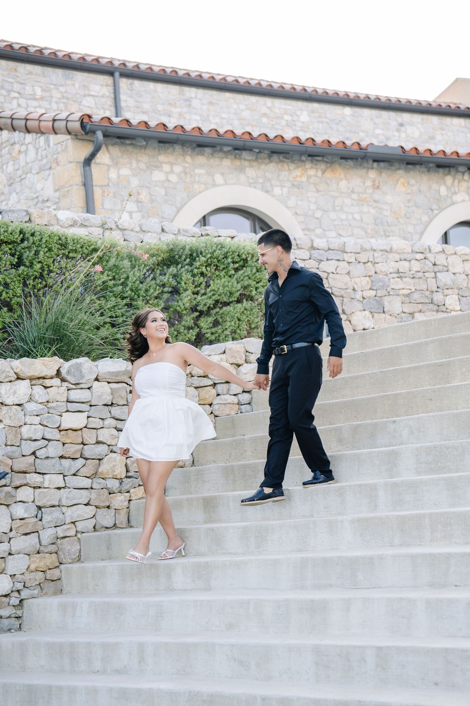Couple holding hands on stone steps outside a rustic building, woman in white dress and man in black outfit.