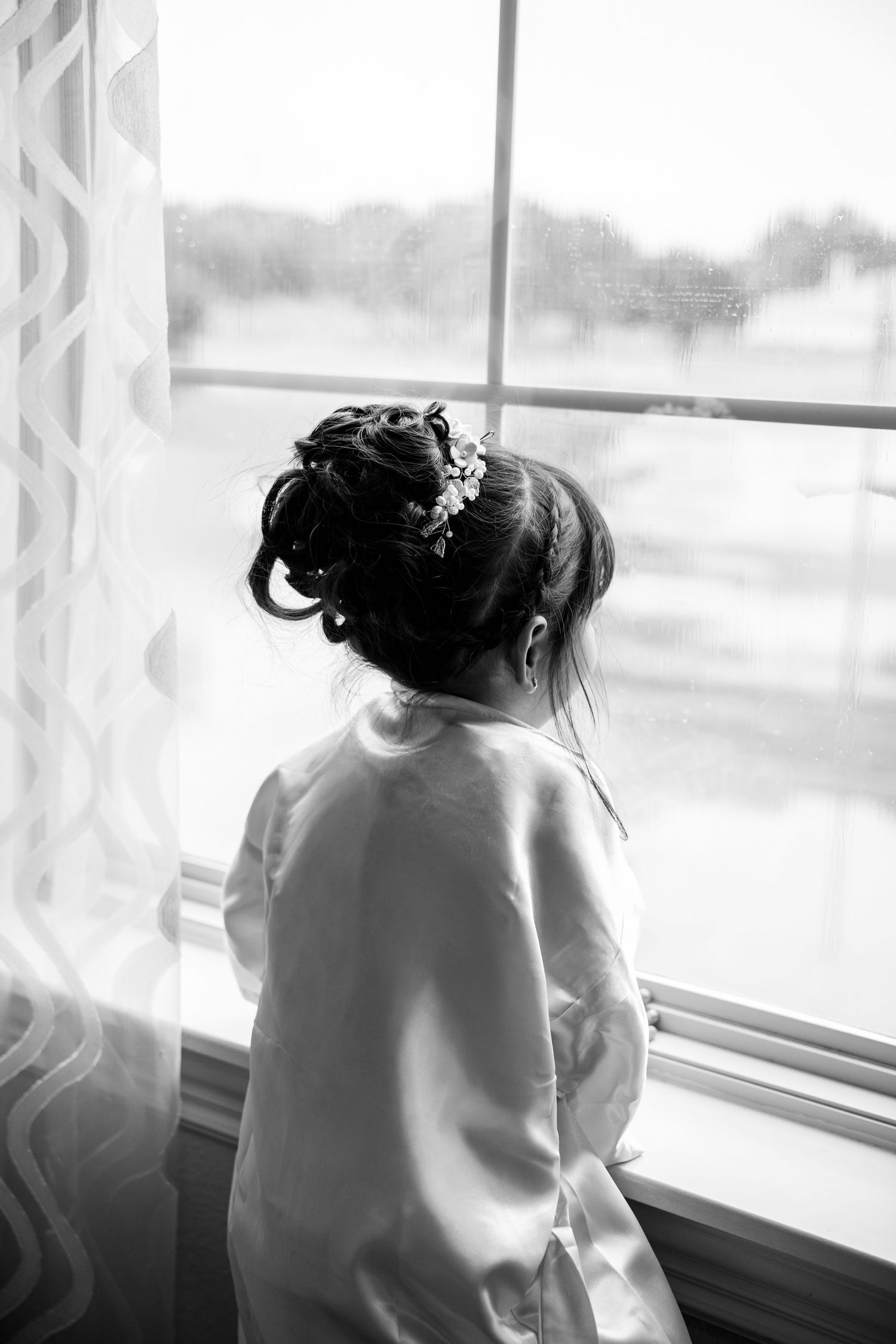 Black and white photo of a little girl in a robe with an elegant updo and floral hair accessory gazing out a window. Burleson Tx