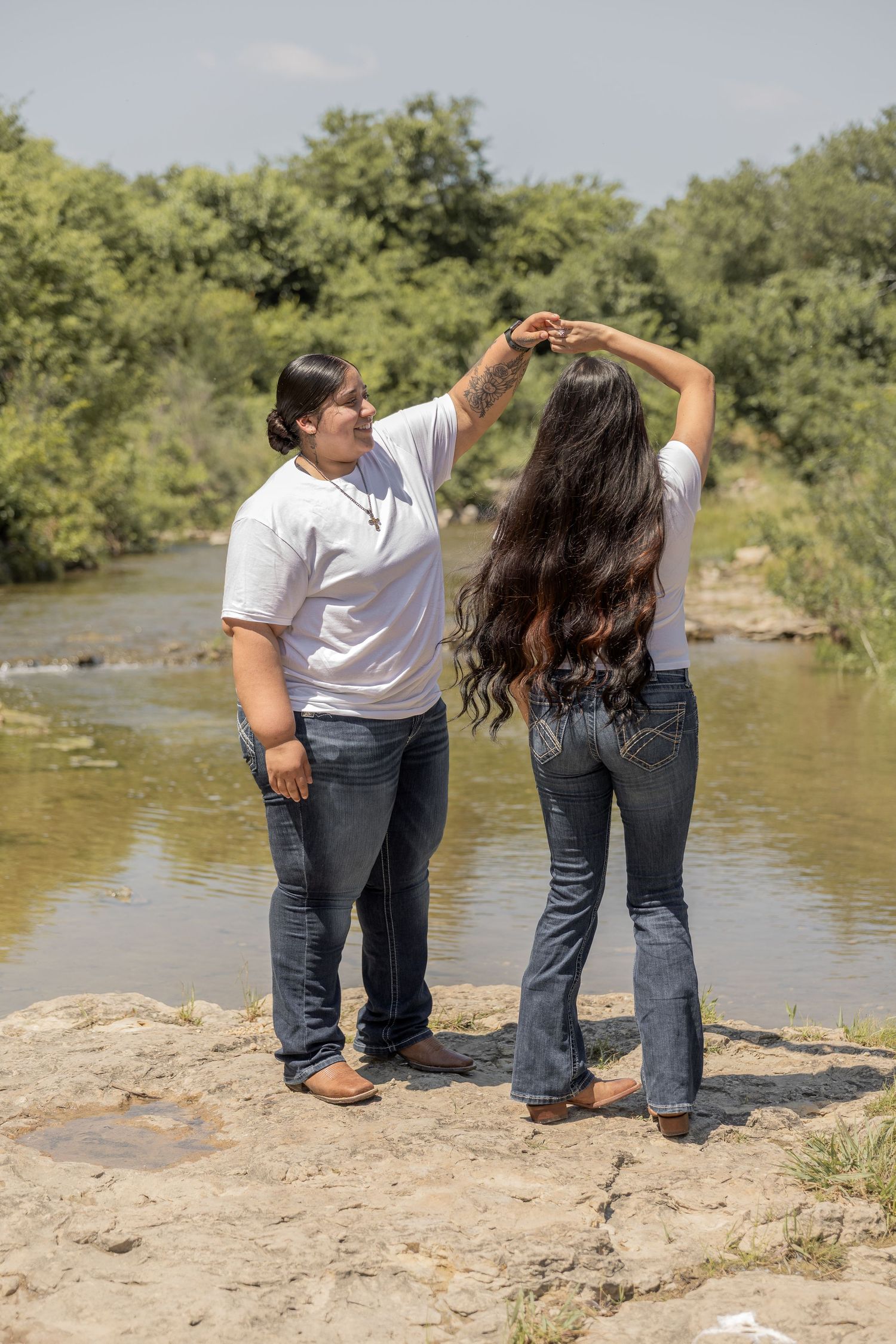 A couple dances outdoors near a river, man spinning woman with long dark hair in jeans and white tops.
