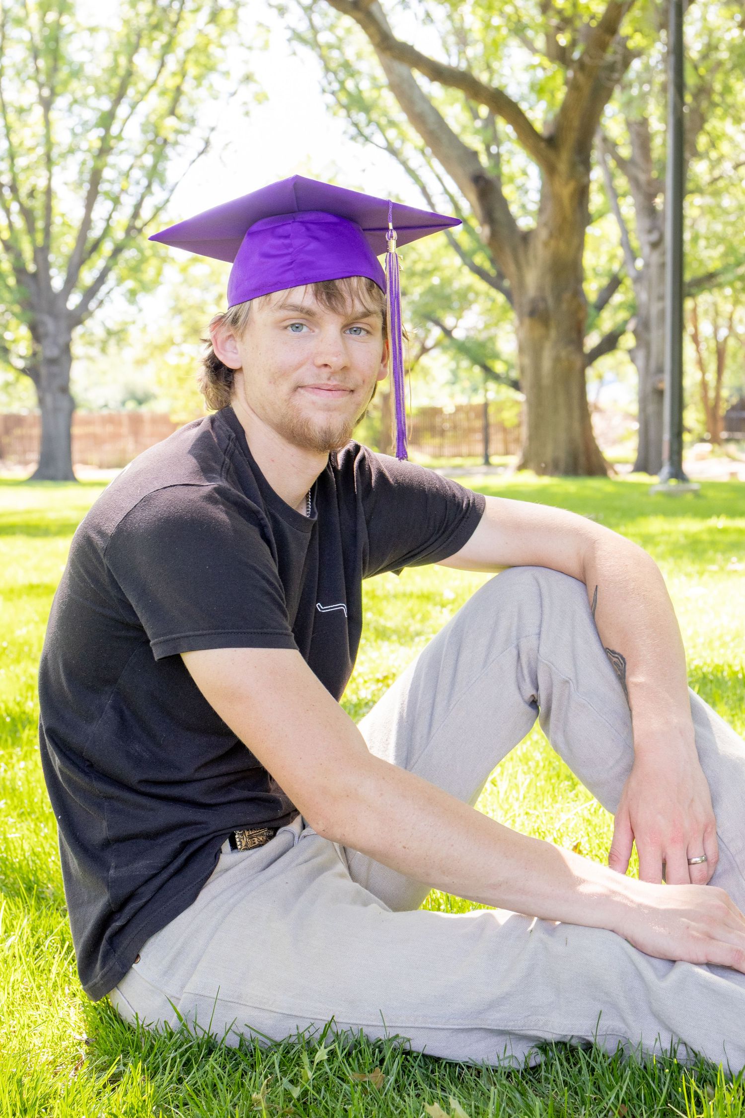 Young man in black shirt and purple graduation cap sits cross-legged on lush green grass.