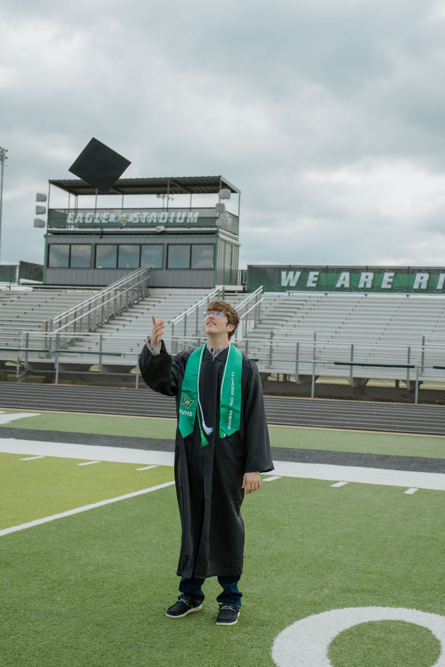 High school graduate in black cap and gown pointing skyward at Eagle Stadium football field.