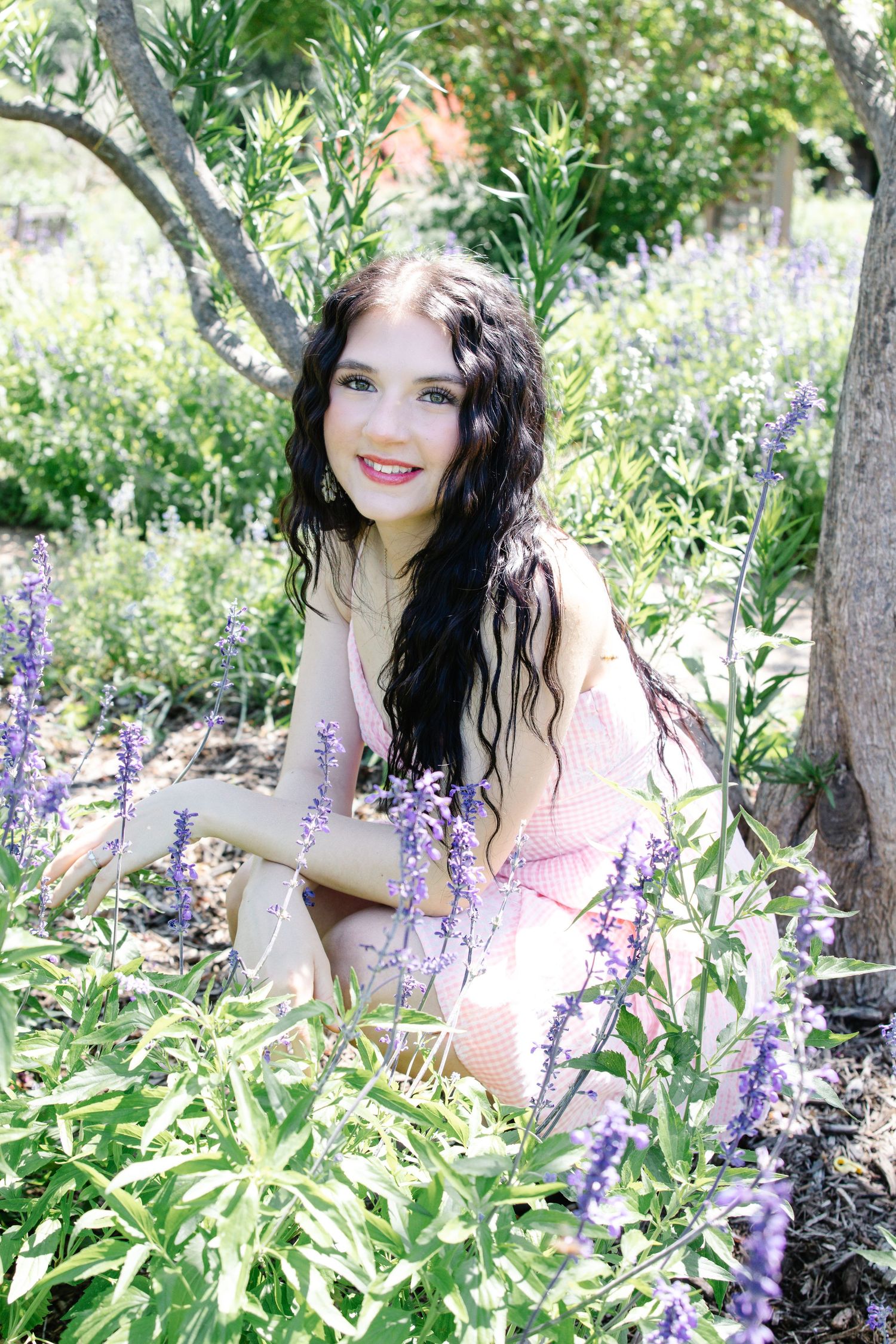 A young woman in a pink dress smiles while sitting among purple lavender flowers in a garden.