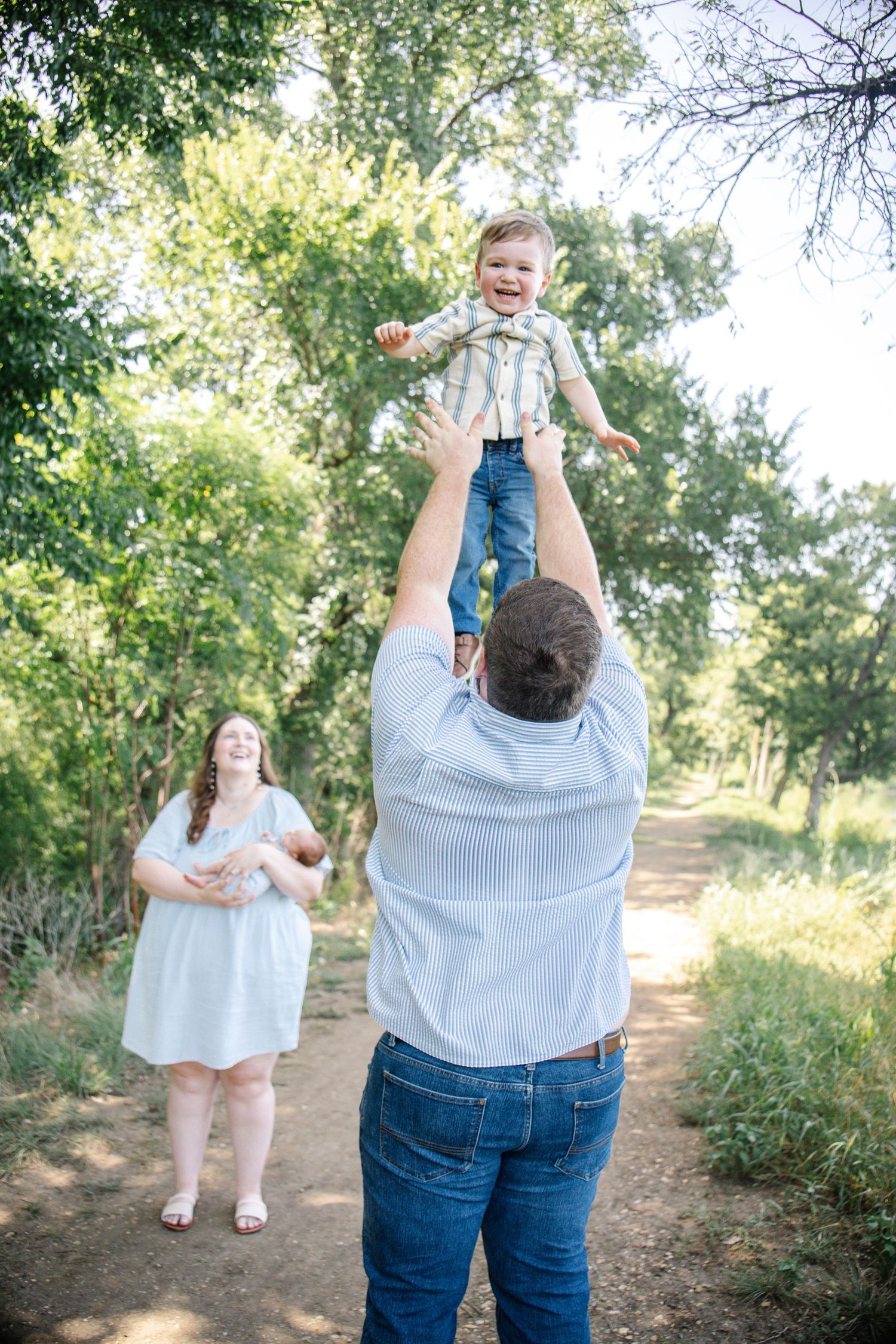 A father joyfully tosses his laughing toddler in the air while mother watches on a wooded path.