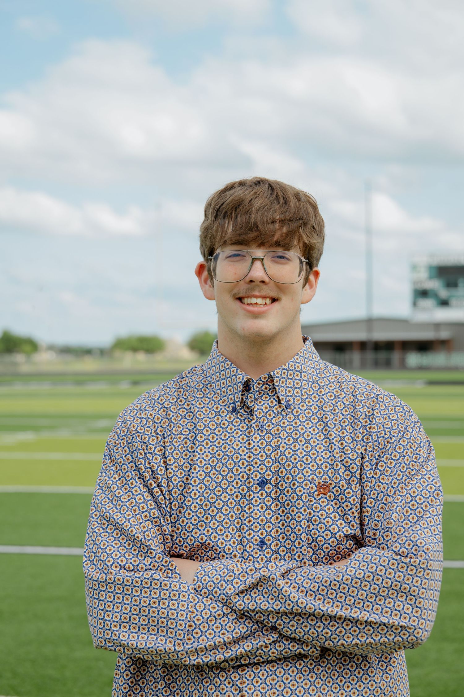Smiling teen boy in patterned shirt with arms crossed standing on a green athletic field outdoors.