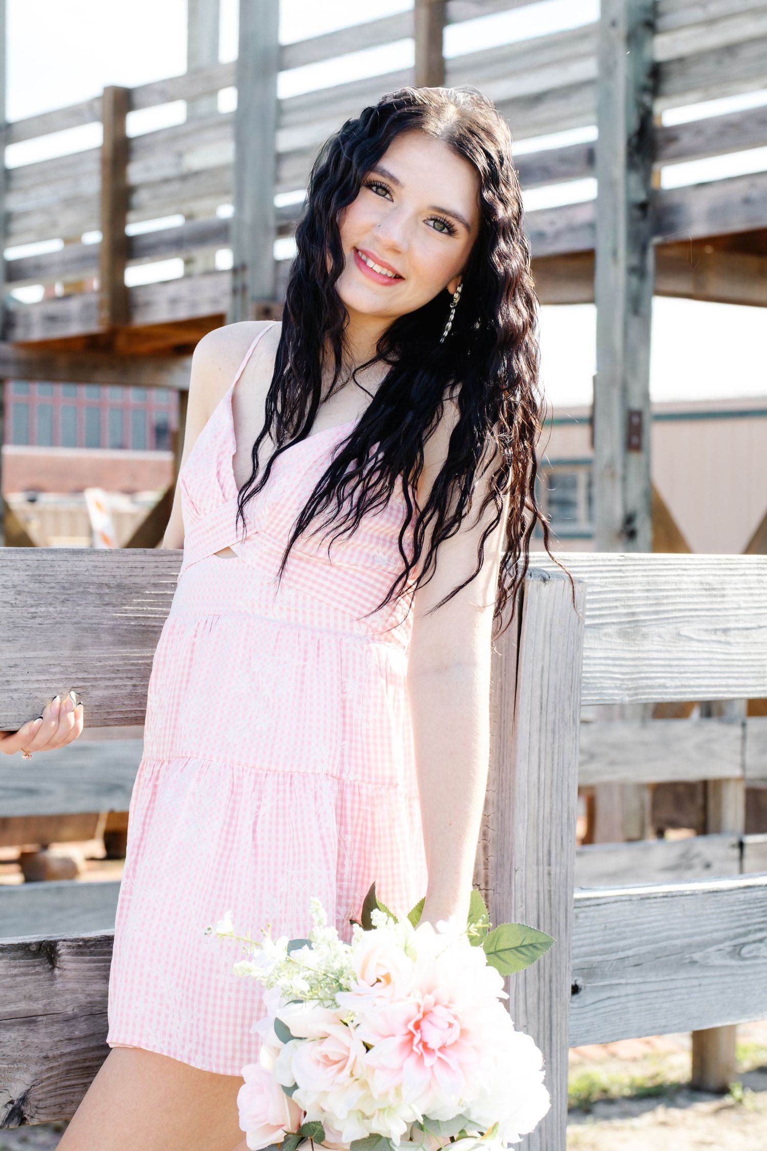 Smiling young woman in pink gingham dress holding floral bouquet near rustic wooden fence.