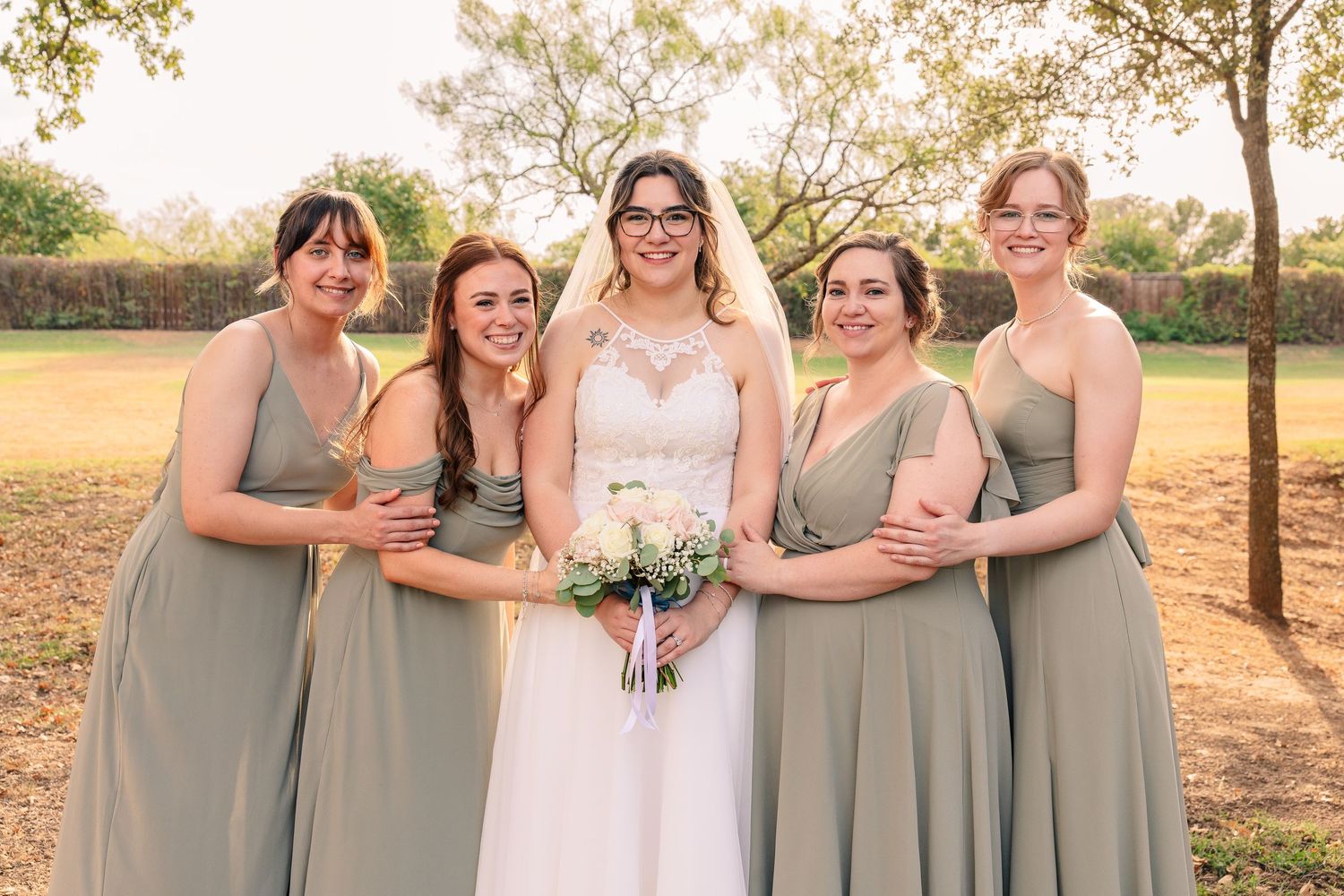 Bride in a white lace gown holding a bouquet poses outdoors with four smiling bridesmaids in matching sage green dresses.