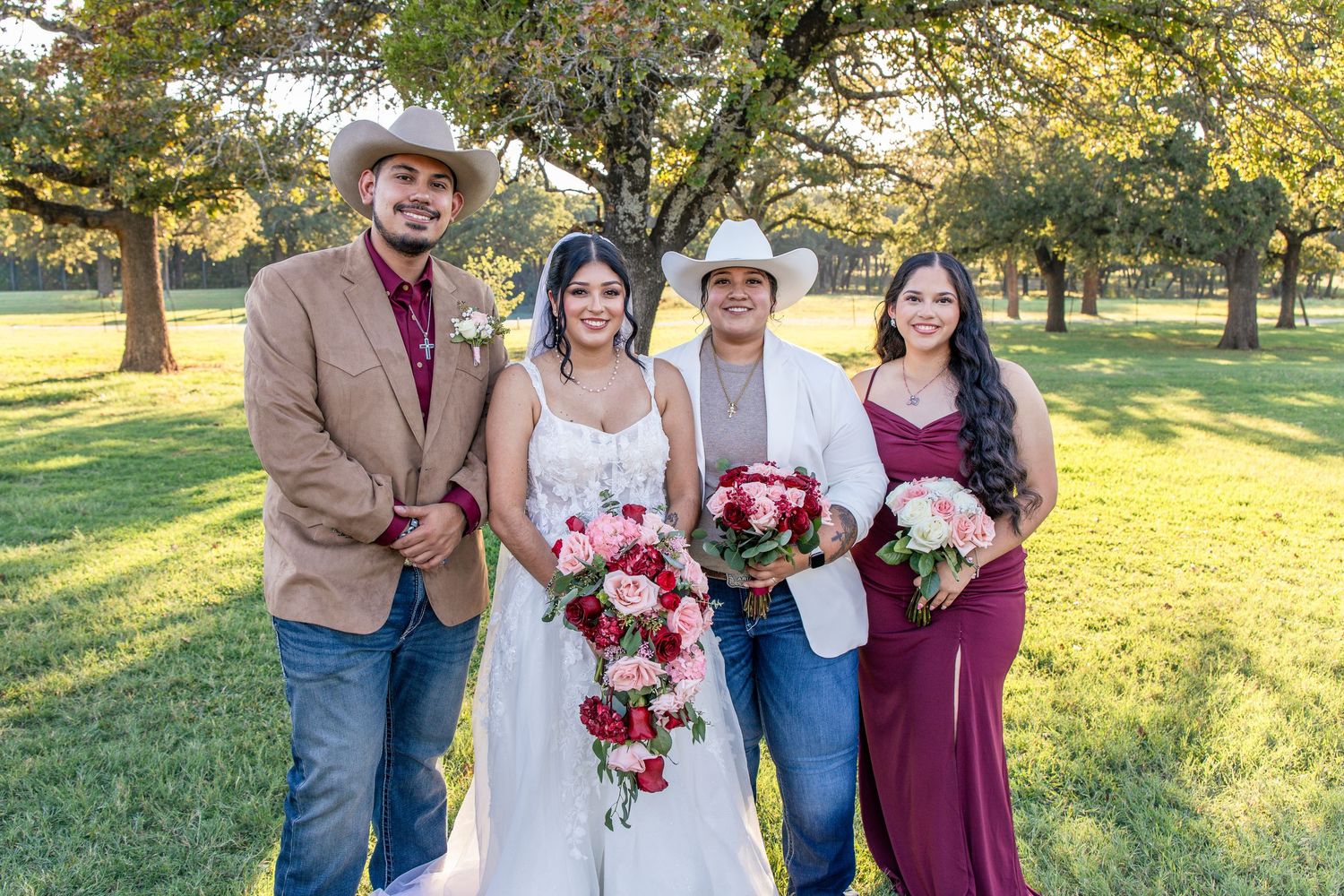 Bride in a white gown holding a red rose bouquet poses outdoors with three wedding party members in cowboy hats under oak trees.