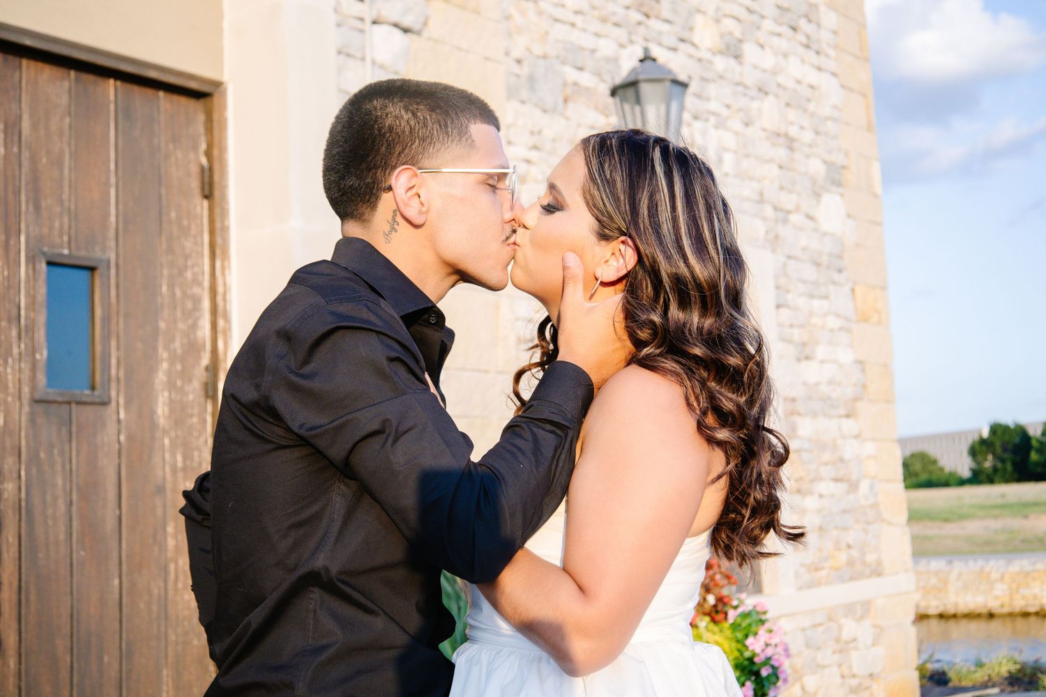 A bride and groom share a romantic kiss outdoors near a stone building on their wedding day.