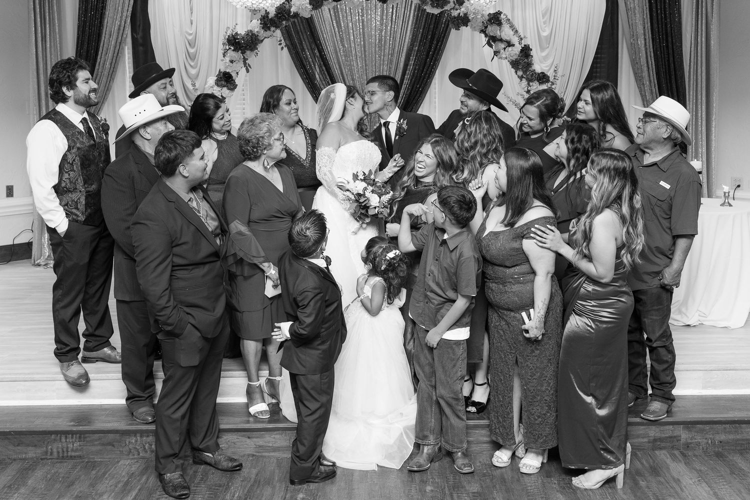 Bride and groom surrounded by joyful wedding party guests in black and white celebration photo.