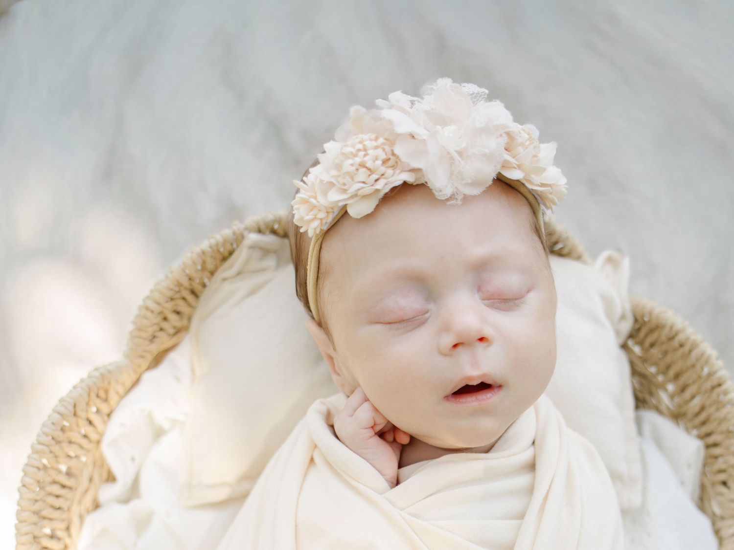 Sleeping newborn baby wrapped in cream swaddle wearing floral headband resting in wicker basket.