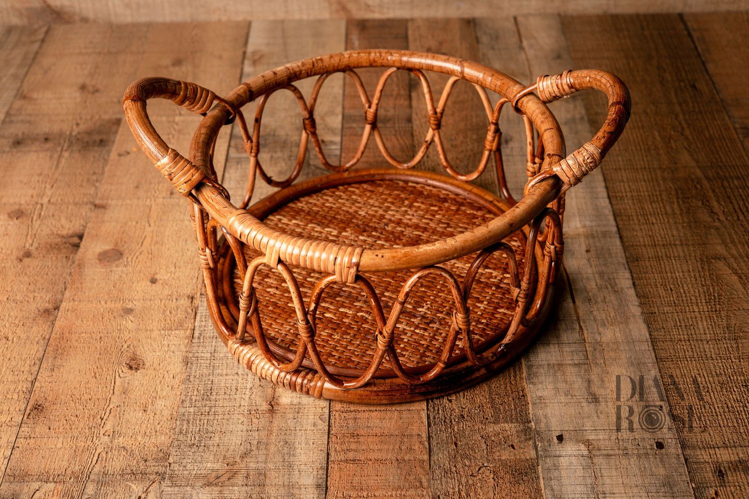 Handwoven rattan wicker basket with looped handles on rustic wooden floor, perfect for newborn photography props.