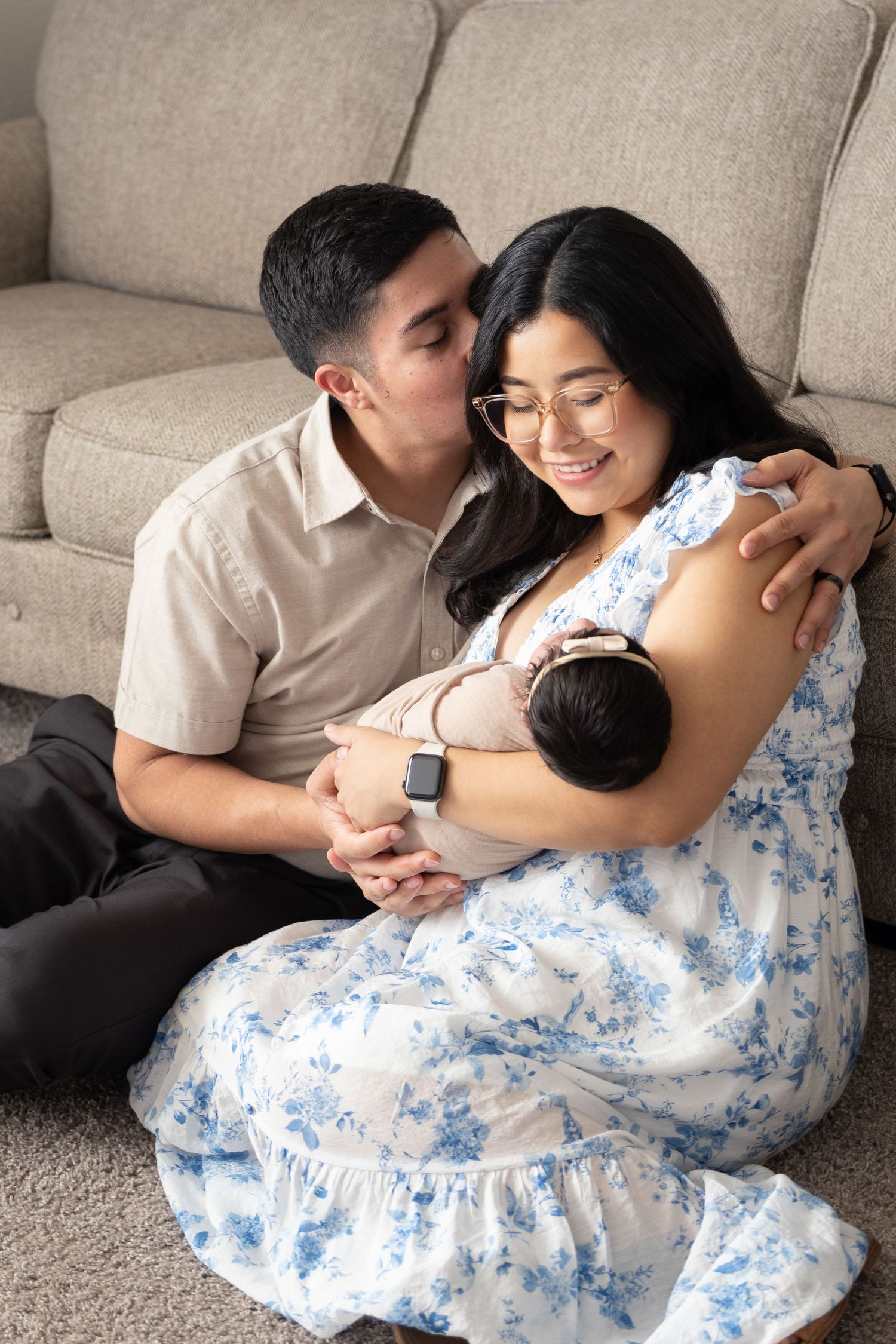 A couple sits on the floor holding their newborn baby, the man kissing the woman's cheek as she smiles warmly.