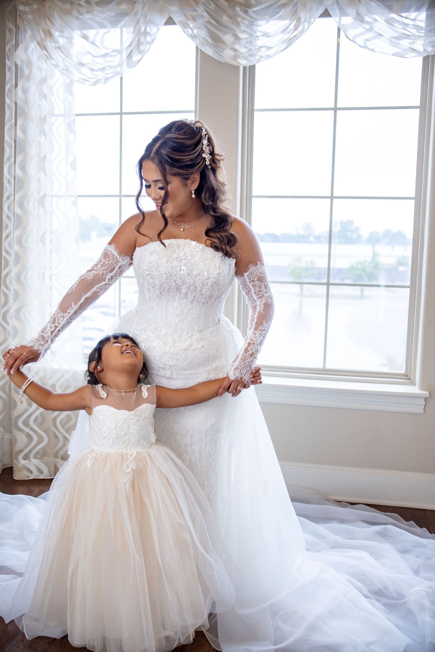 Bride in lace wedding gown stands with young flower girl by sunlit window.