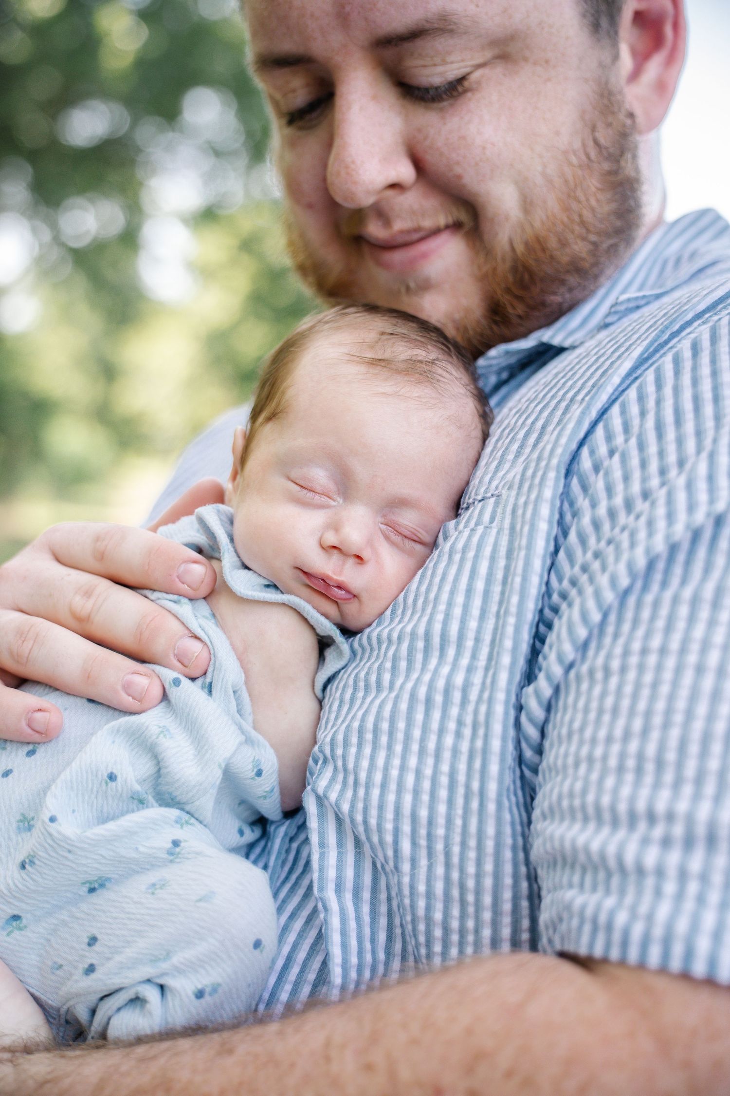 Bearded father smiling while cradling his sleeping newborn baby dressed in blue outdoors.