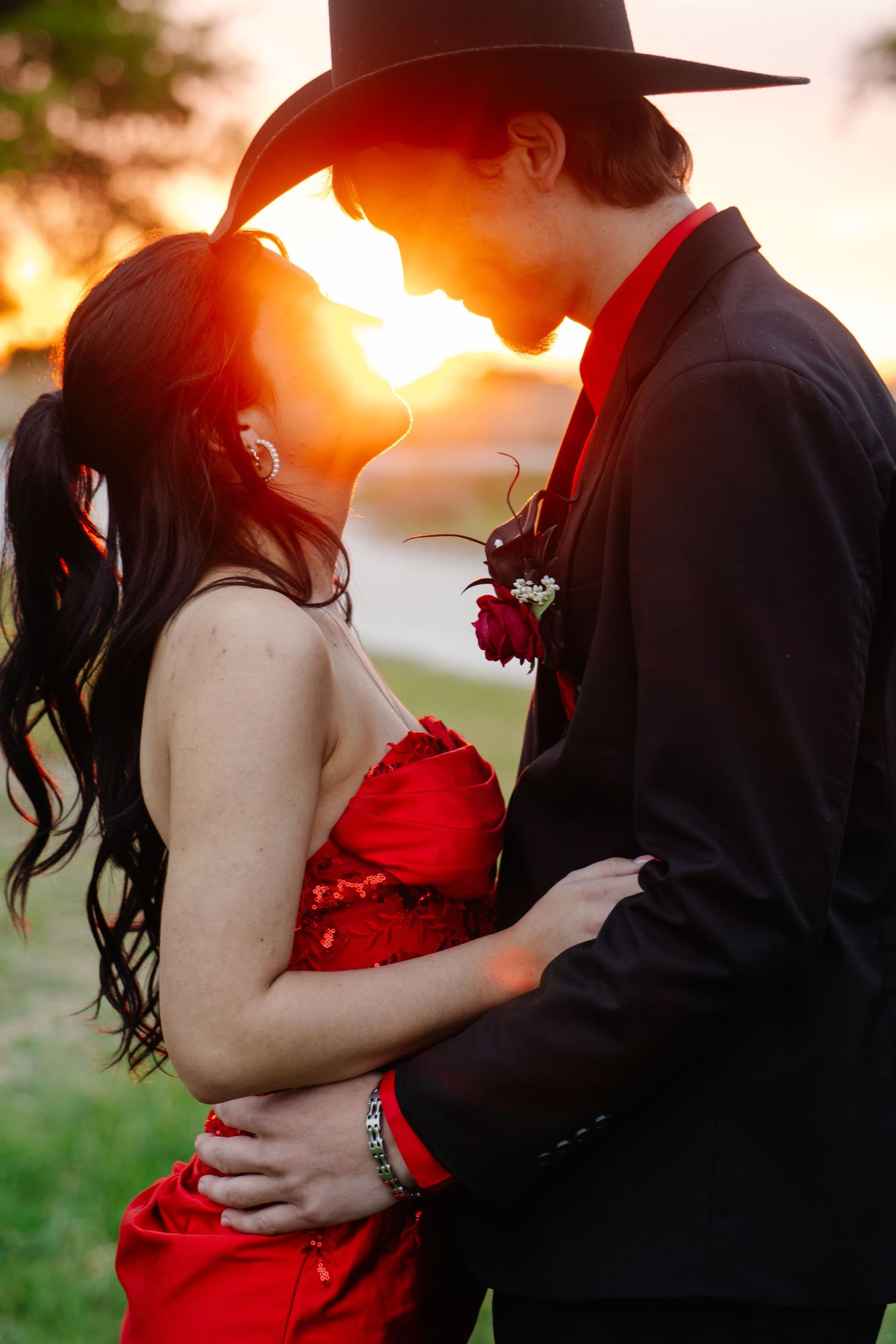 Romantic prom couple nearly kissing at sunset, girl in red sequin dress, boy in black cowboy hat.