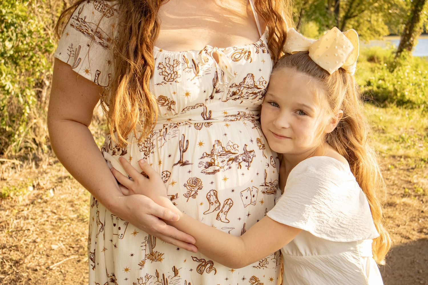 Young girl hugging a pregnant woman's baby bump outdoors in warm golden sunlight during a maternity photo session.