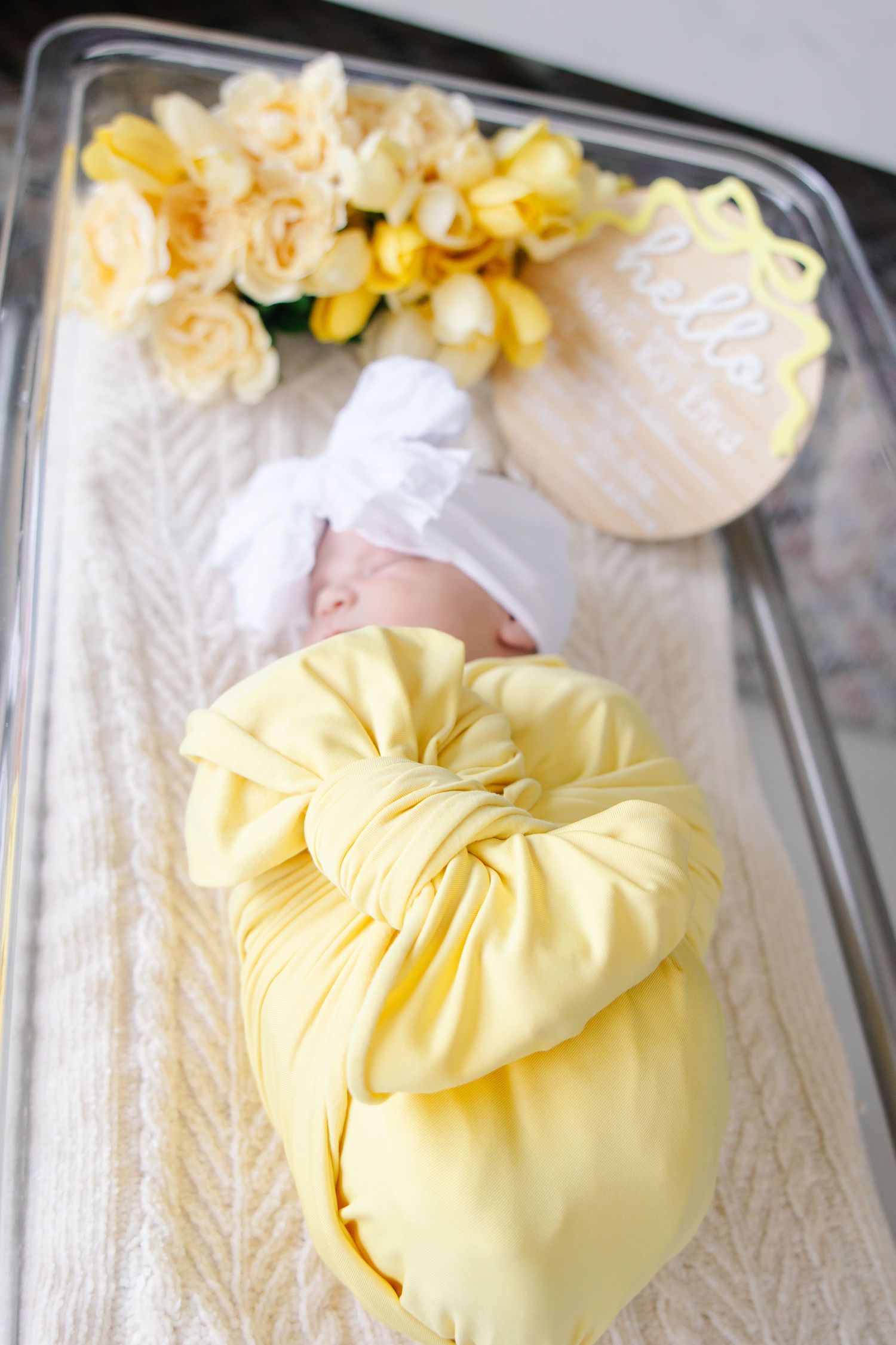 Newborn baby in yellow swaddle with white bow headband in hospital bassinet with yellow flowers.