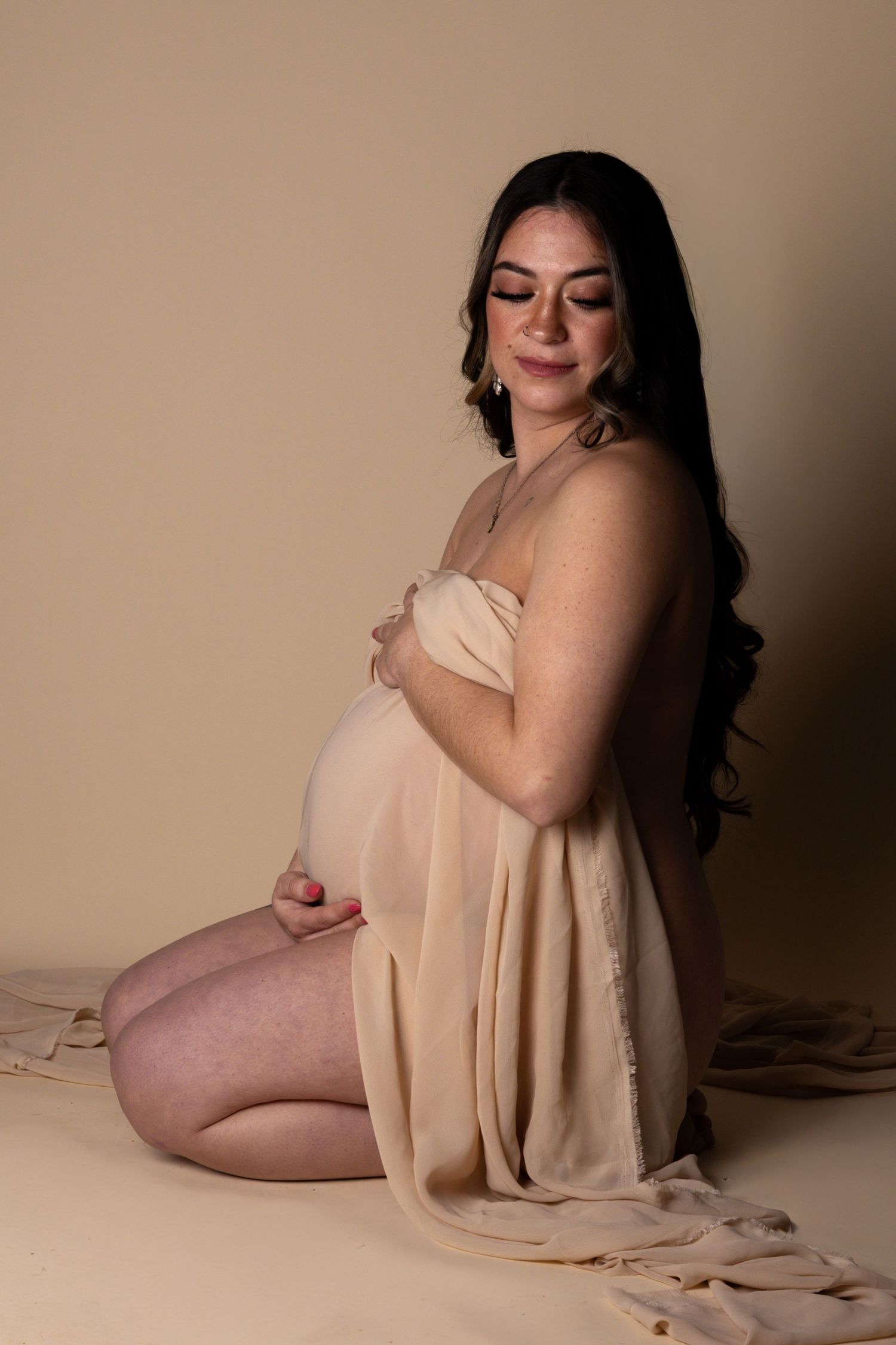 Pregnant woman draped in beige fabric kneeling gracefully in a soft-lit studio maternity portrait.