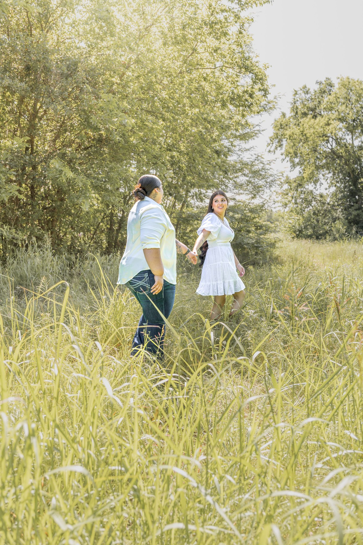 Couple holding hands walking through tall grass in a sunlit meadow surrounded by green trees.
