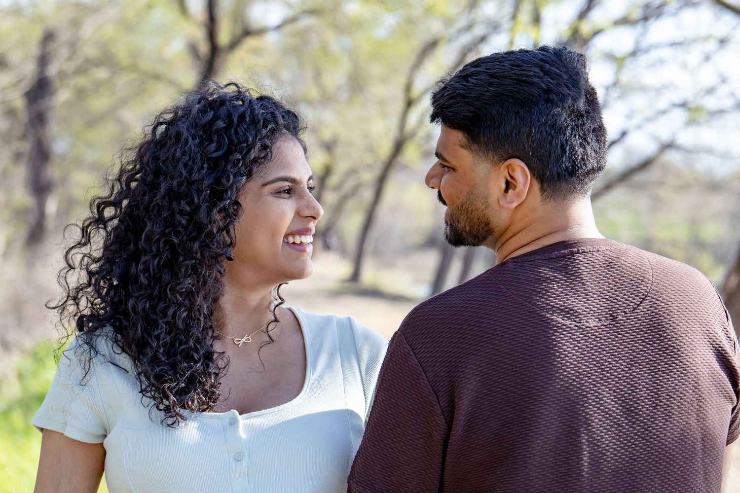 Happy couple smiling at each other outdoors in a sunlit park setting with trees in the background.