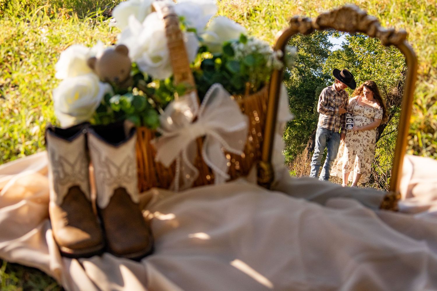 Country-style maternity setup with cowboy boots, white floral bouquet, and couple reflected in ornate gold mirror.