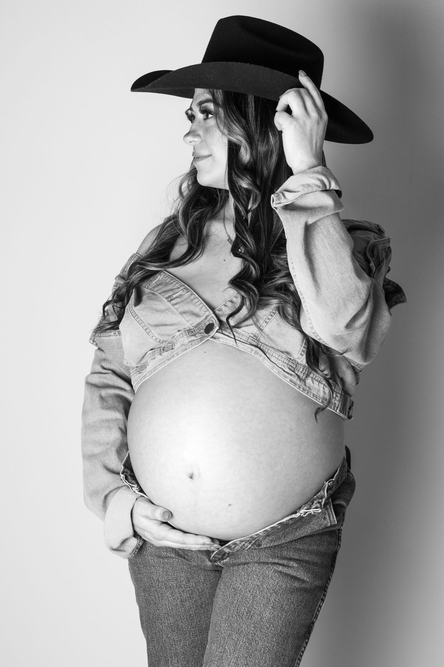Black and white photo of a pregnant woman in a cowboy hat, denim jacket, and jeans cradling her baby bump.