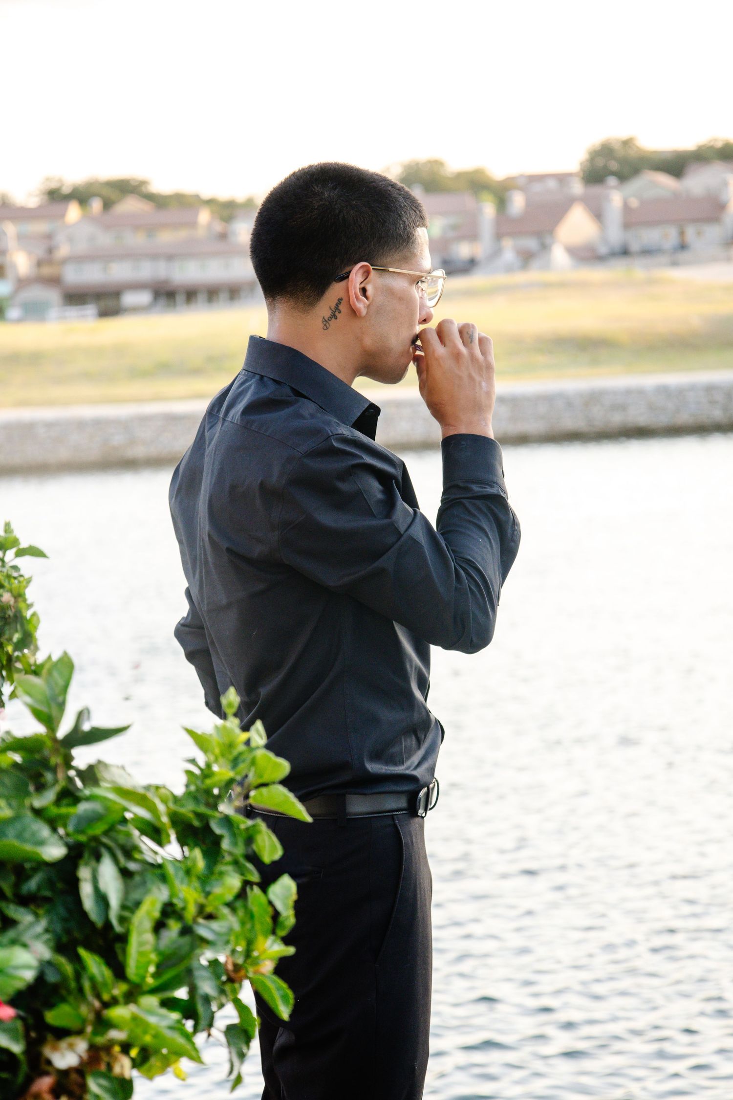 Young man in dark shirt and glasses standing by waterfront, looking thoughtful in profile view.