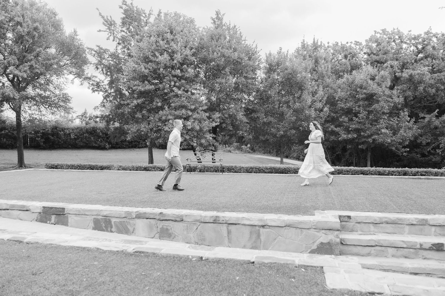 Black and white photo of a bride and groom running playfully across a manicured lawn surrounded by tall trees.