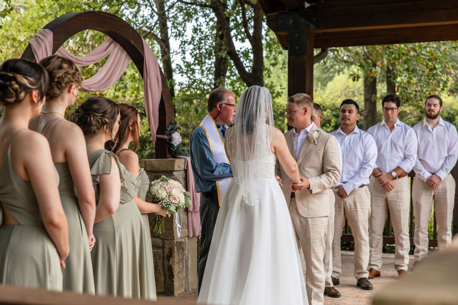 Bride in a white gown and veil holds hands with the groom during an outdoor wedding ceremony as bridesmaids and groomsmen look on.