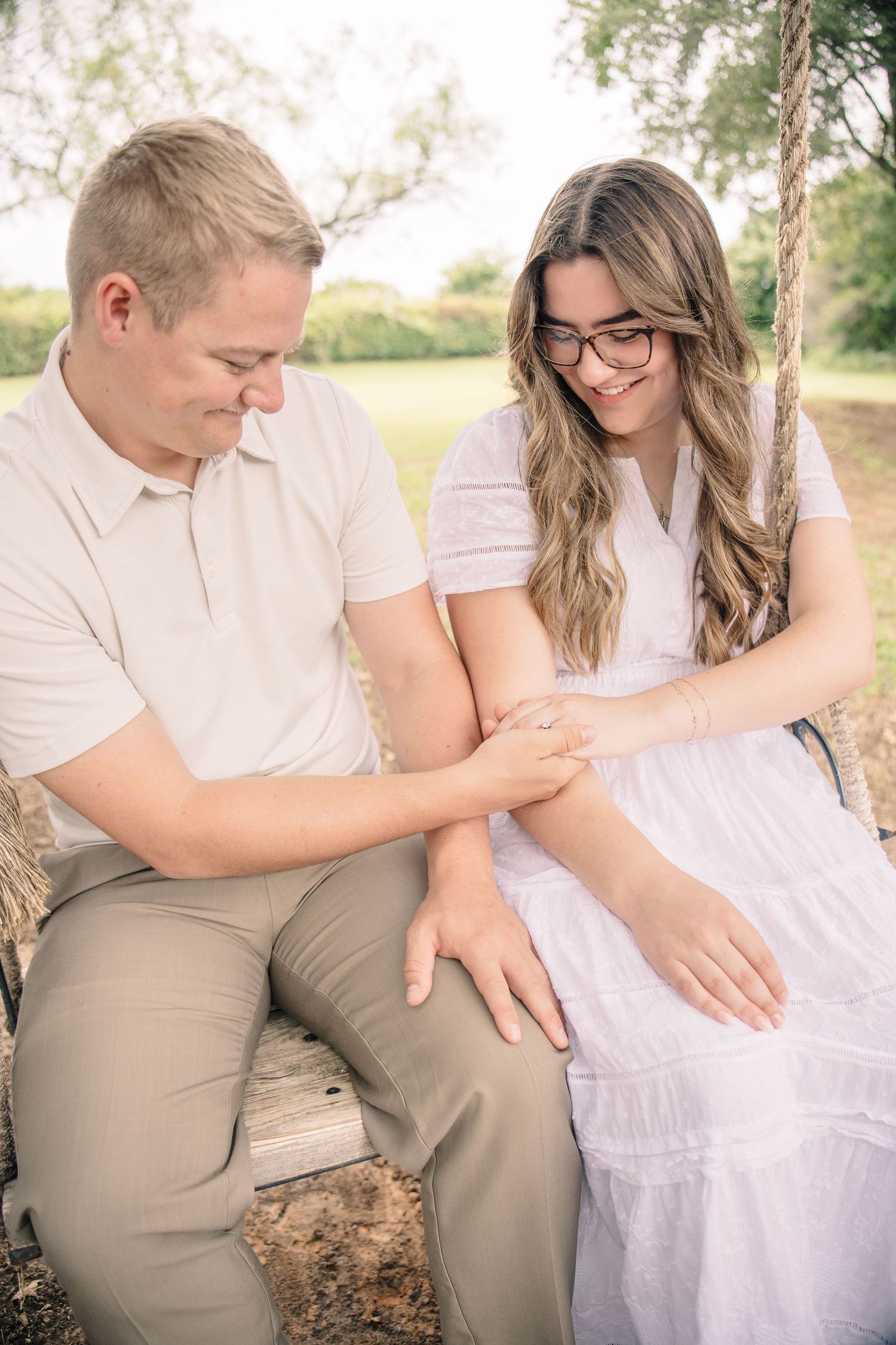 Couple sitting on wooden swing holding hands, smiling at engagement ring outdoors.