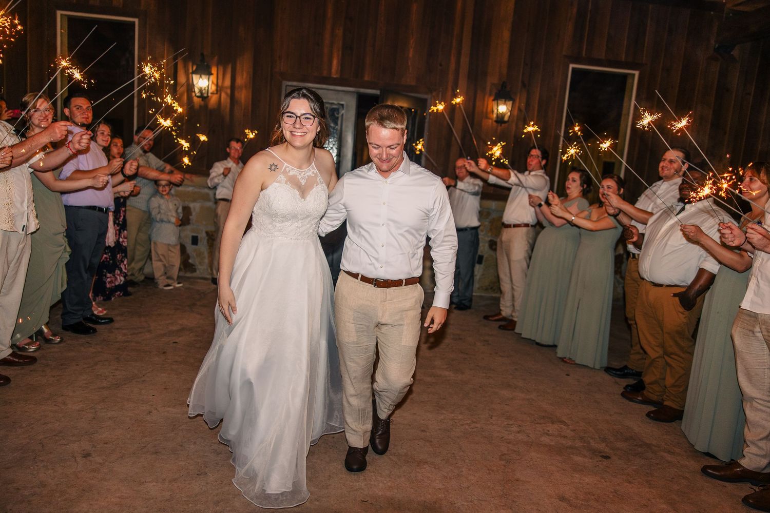 Bride and groom walk hand in hand through a sparkler send-off as wedding guests line the path in a rustic barn venue.