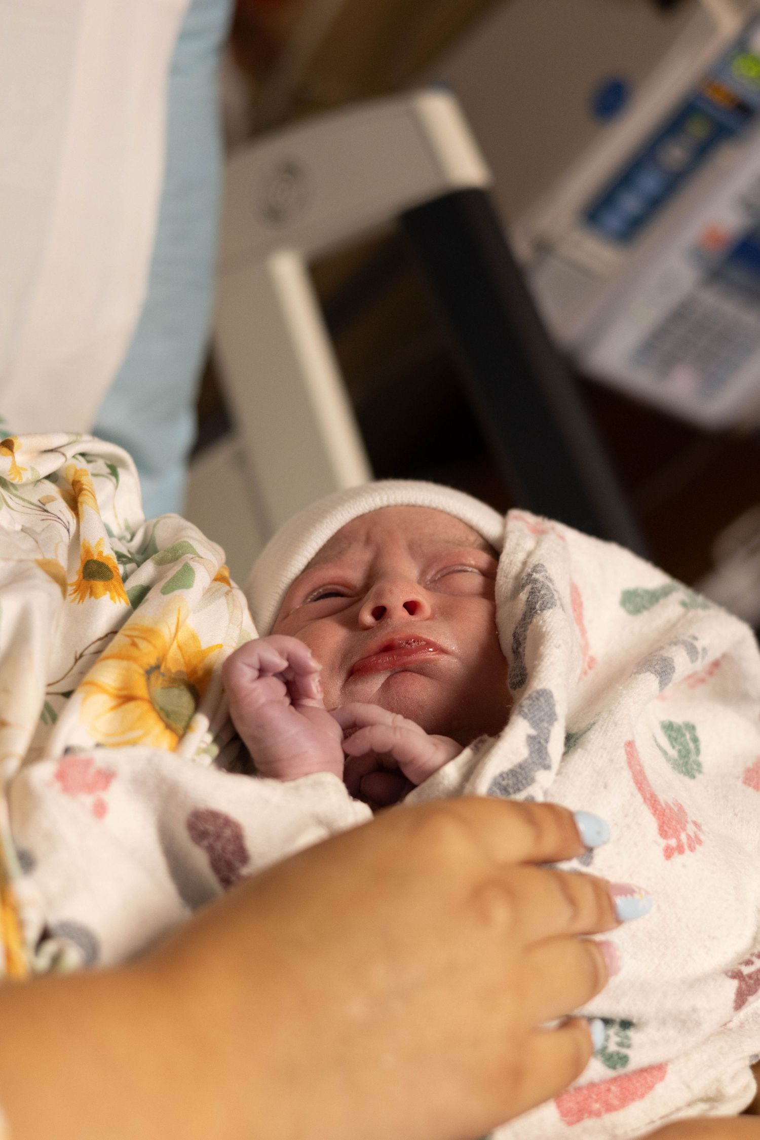 Newborn baby swaddled in floral blanket with tiny fists up, held by mother in hospital.