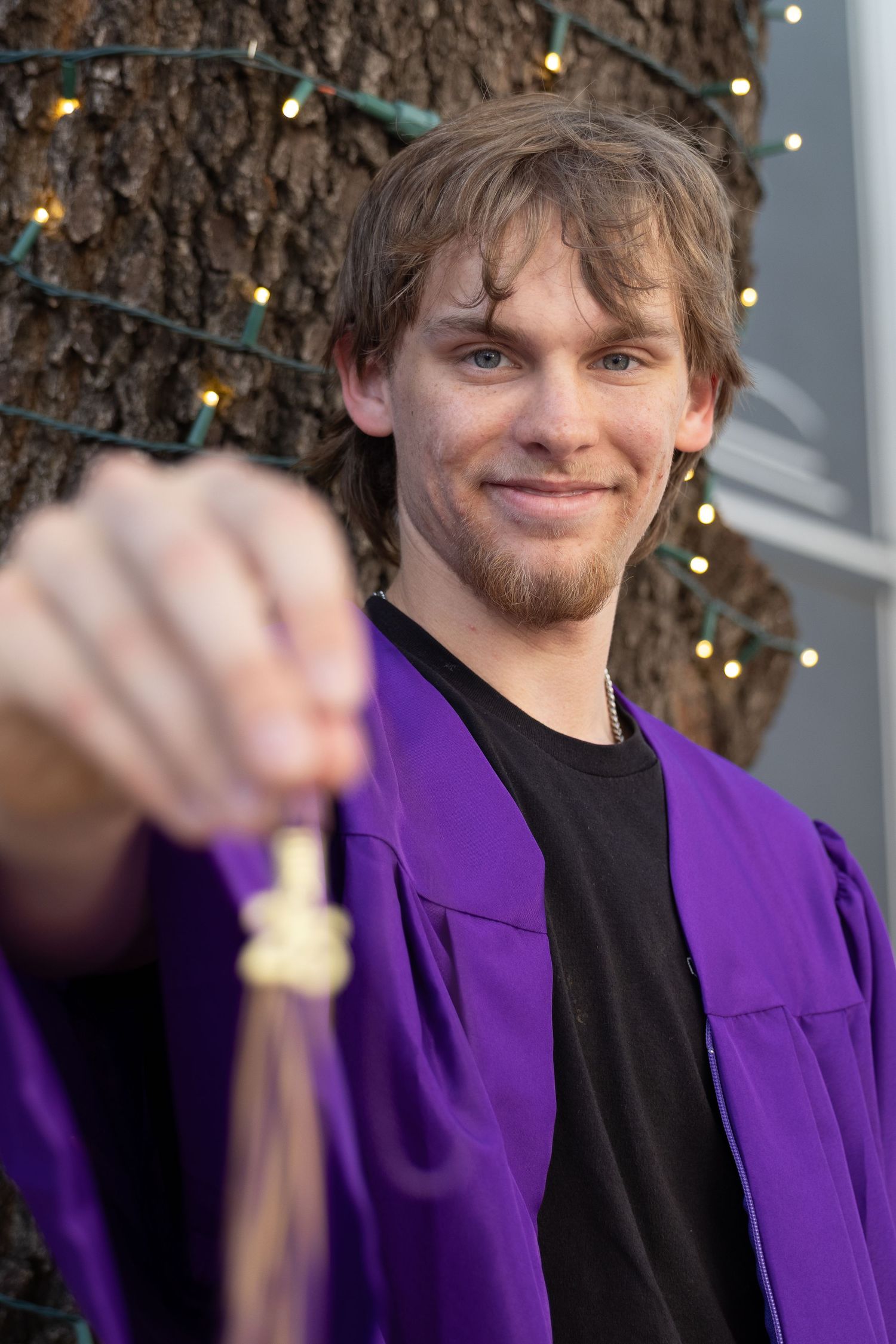 Young man in purple graduation gown smiling while holding tassel toward camera outdoors.