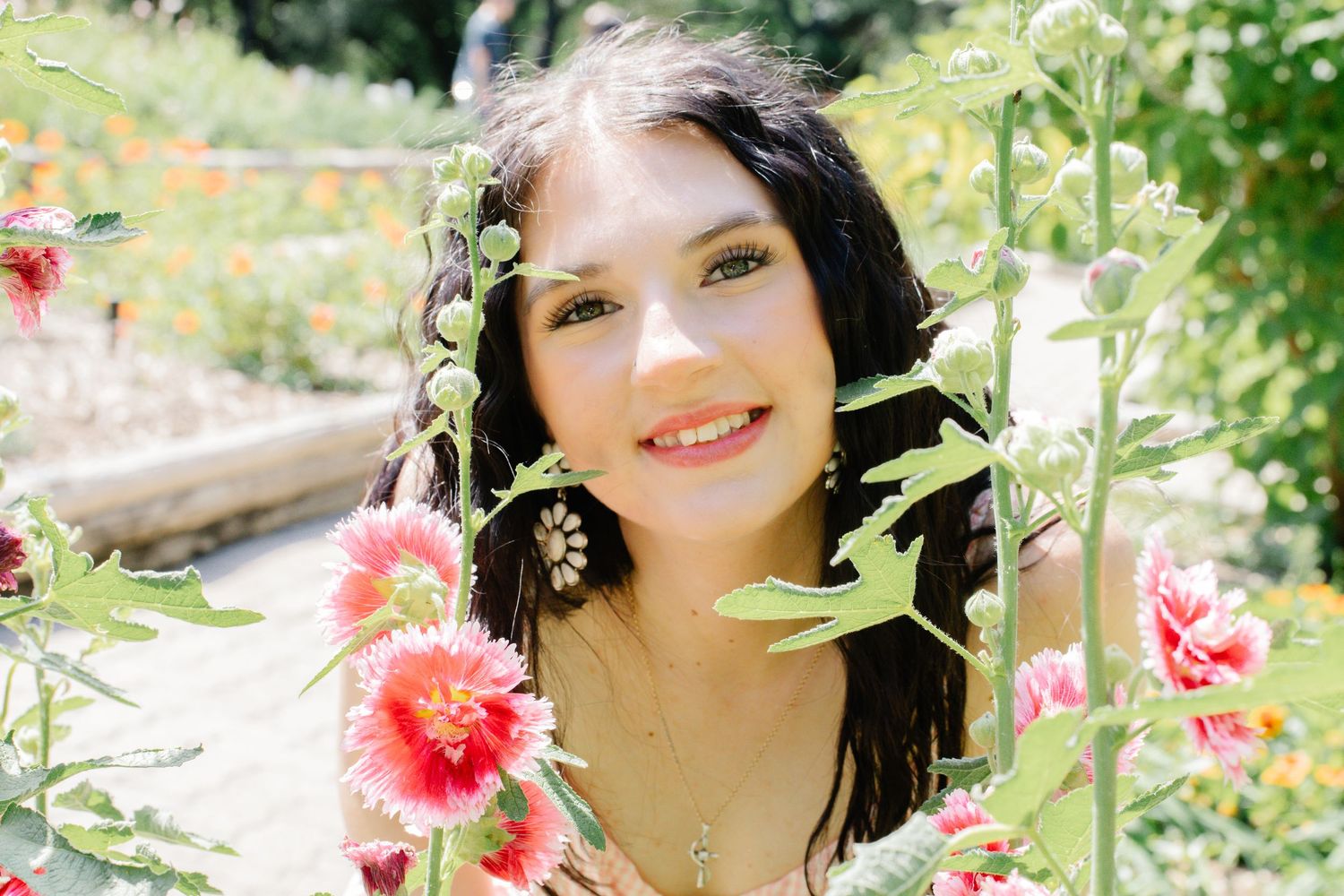 Smiling young woman with dark hair surrounded by blooming pink flowers in a sunny garden.