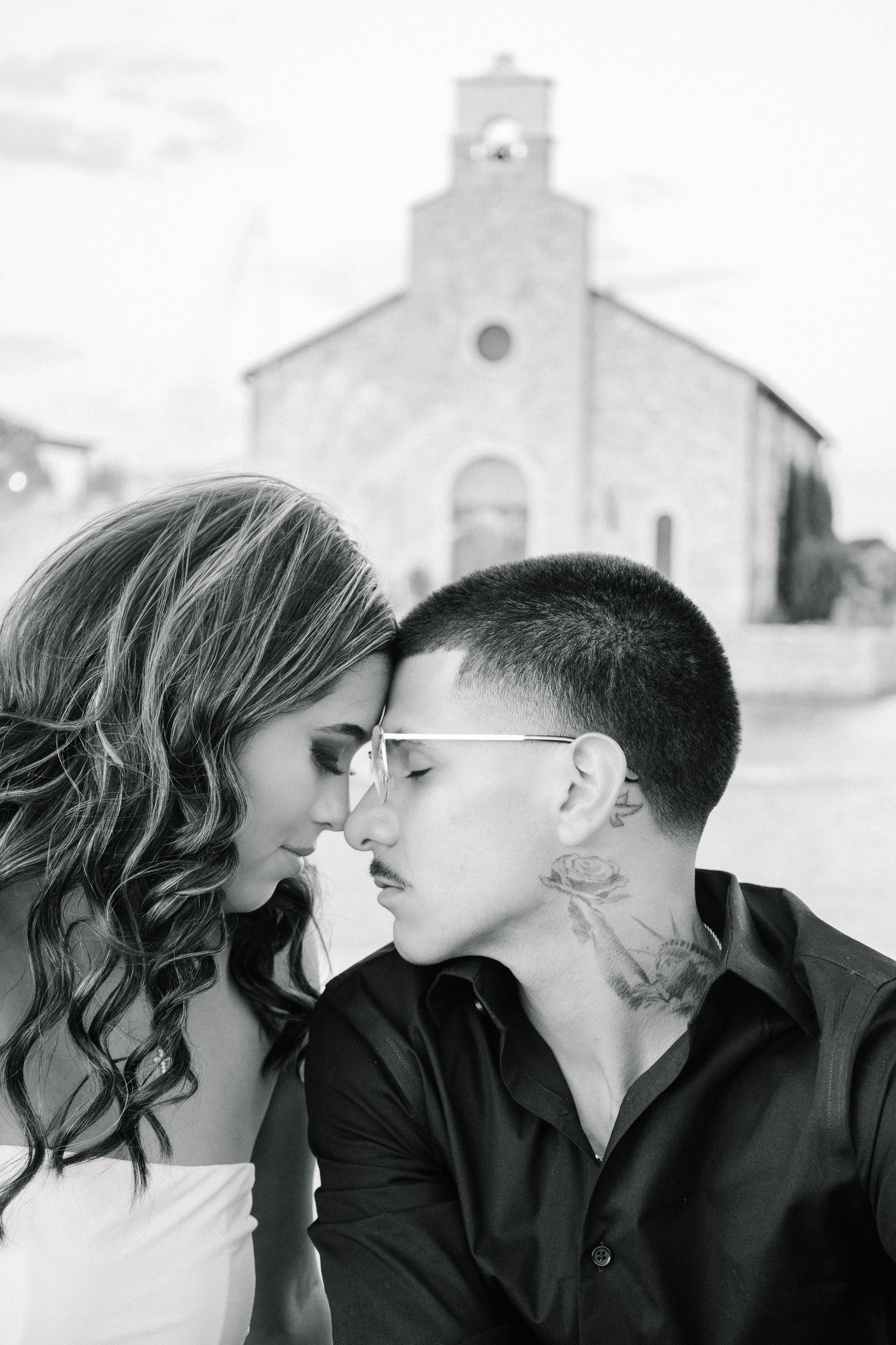 Black and white photo of two people sharing an intimate moment near a historic stone church.
