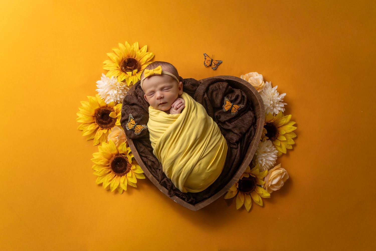 Newborn baby swaddled in yellow wrap sleeping in heart-shaped wooden bowl surrounded by sunflowers on golden background.