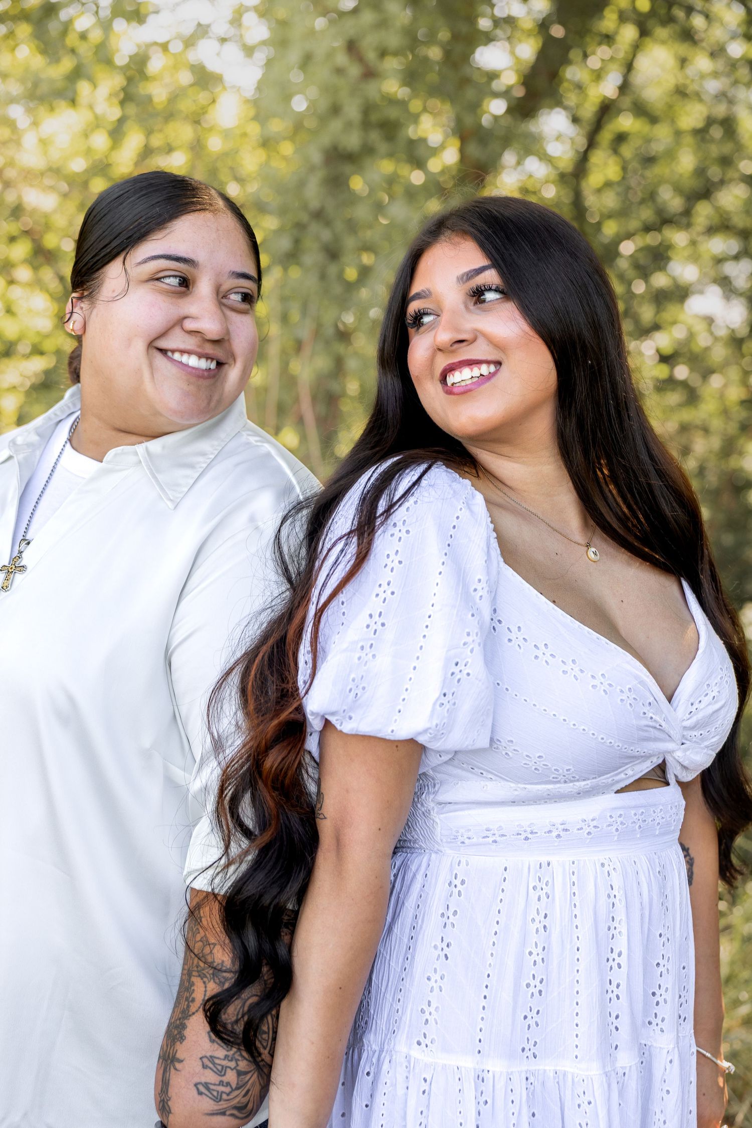 Two smiling women in white outfits posing together in a sunny outdoor park.