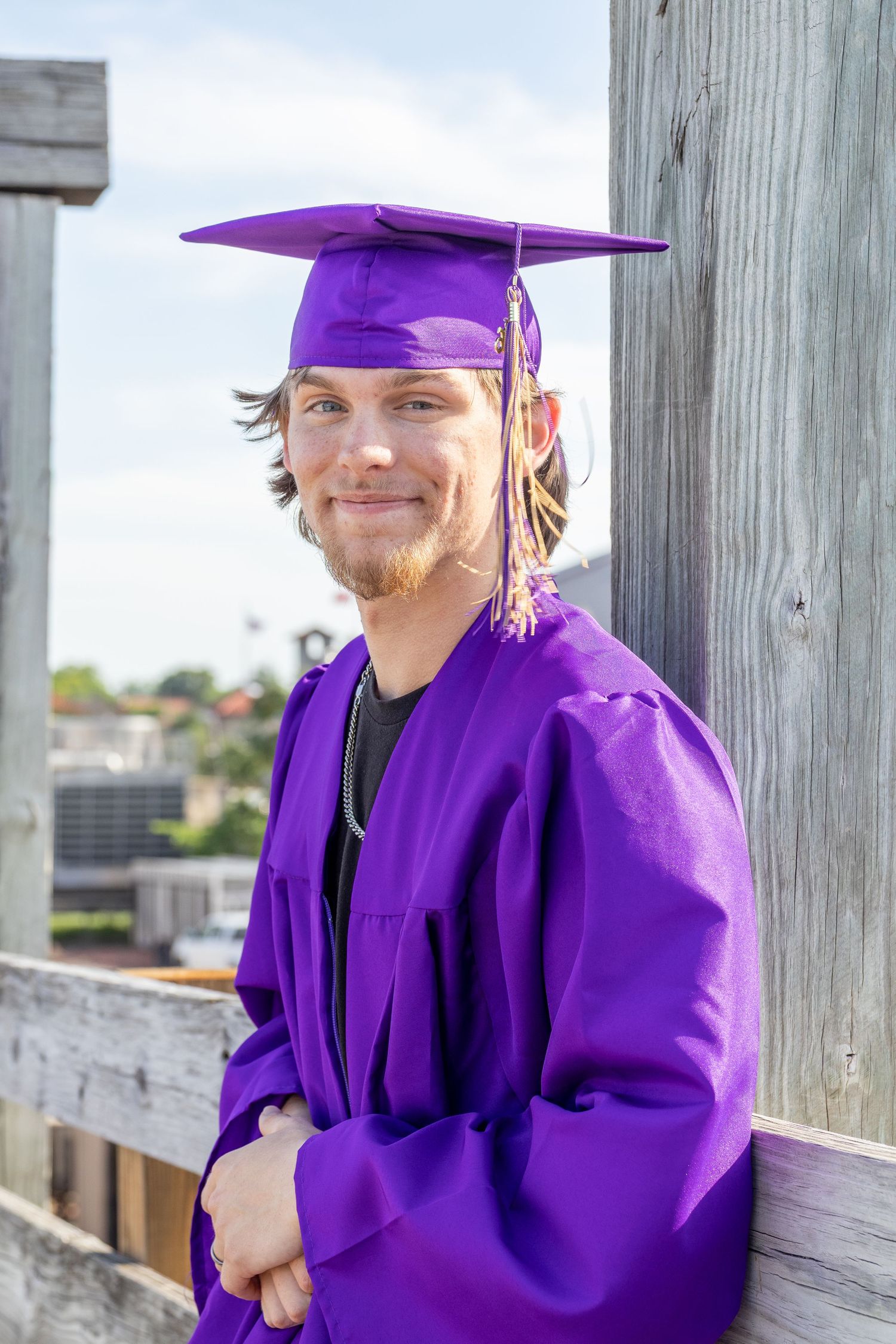 Young male graduate in purple cap and gown leaning against wooden post with city view.