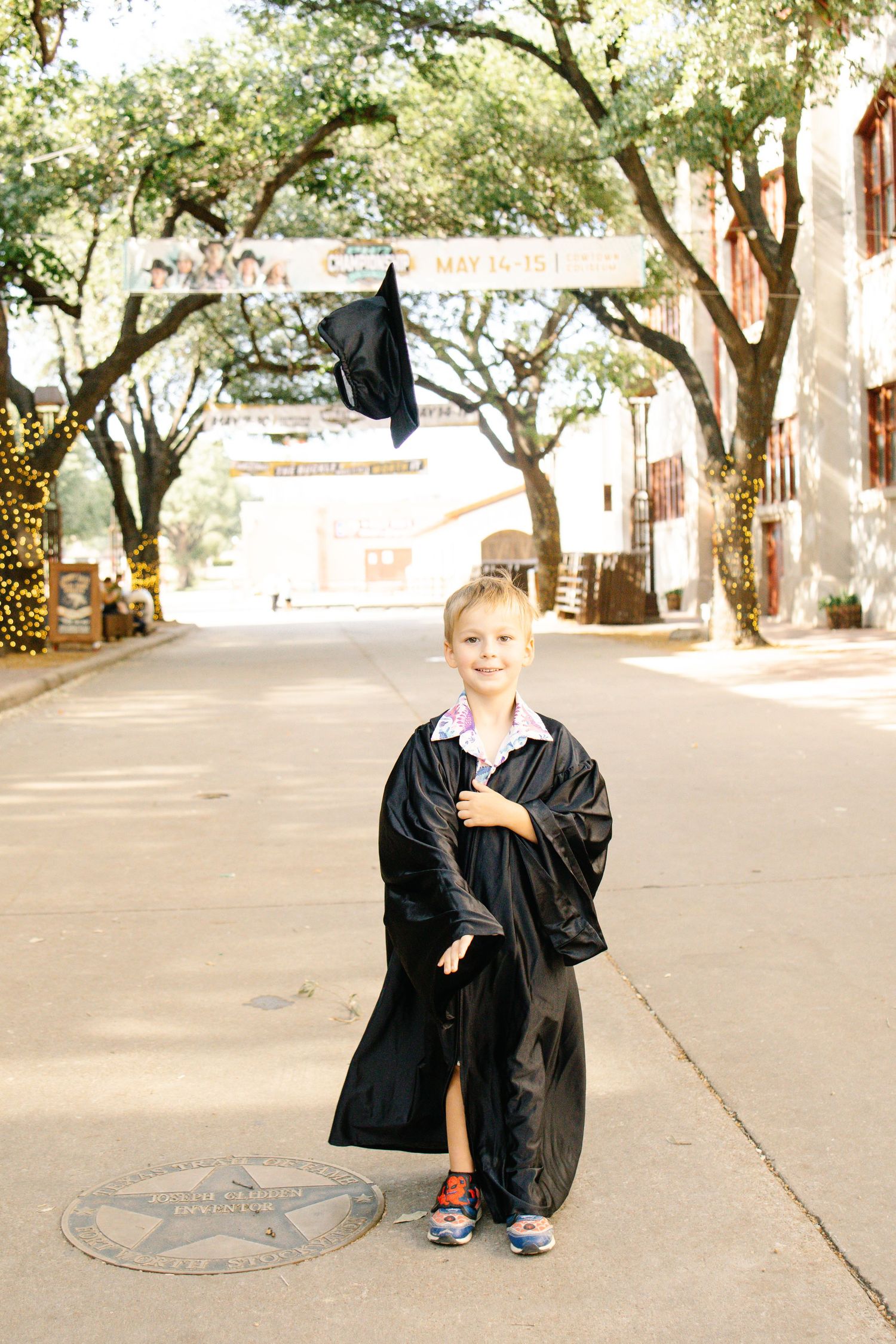 Young blonde boy in black graduation gown and cap standing on a tree-lined street.