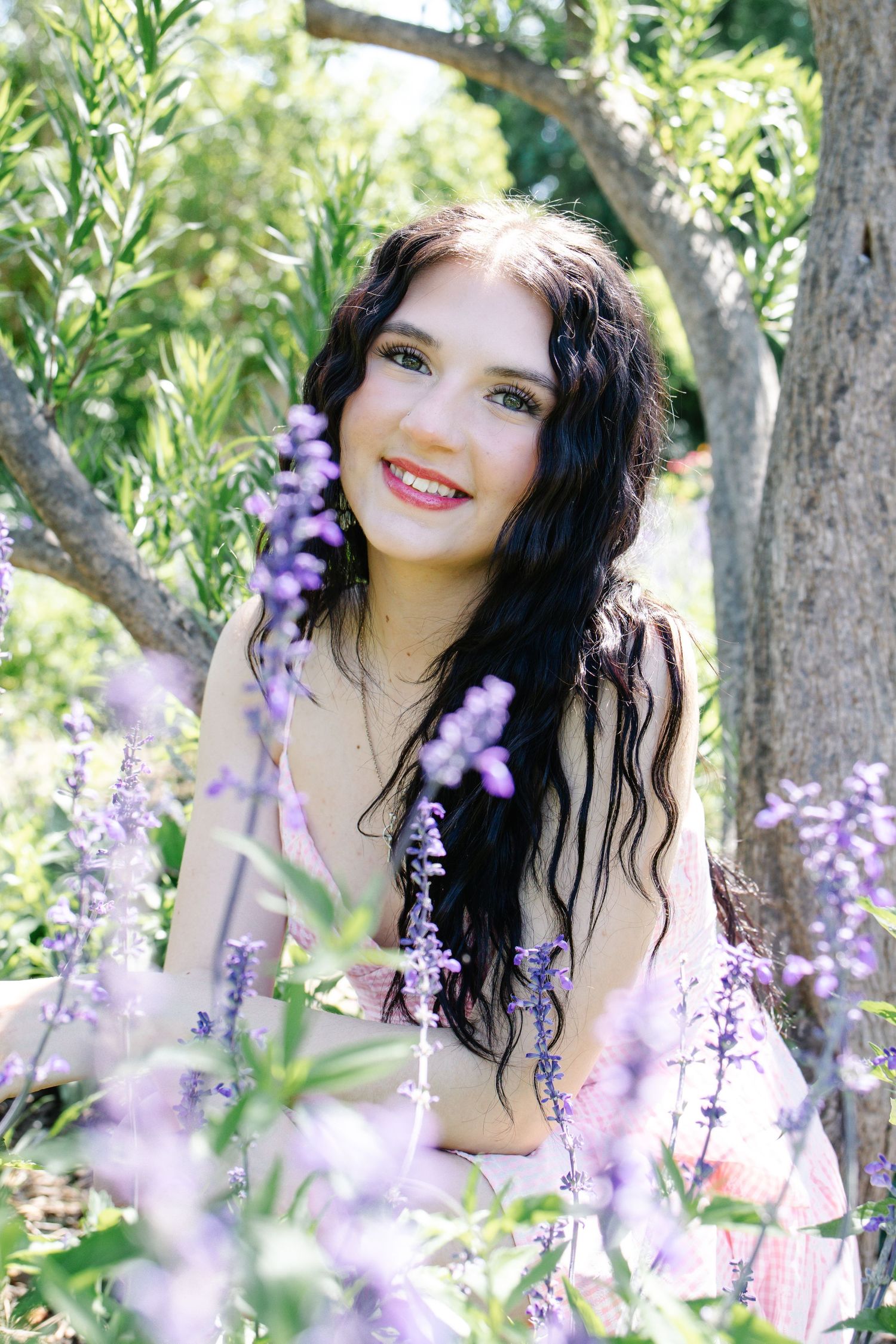 Young woman with dark wavy hair smiling among purple lavender flowers in a sunny garden setting.