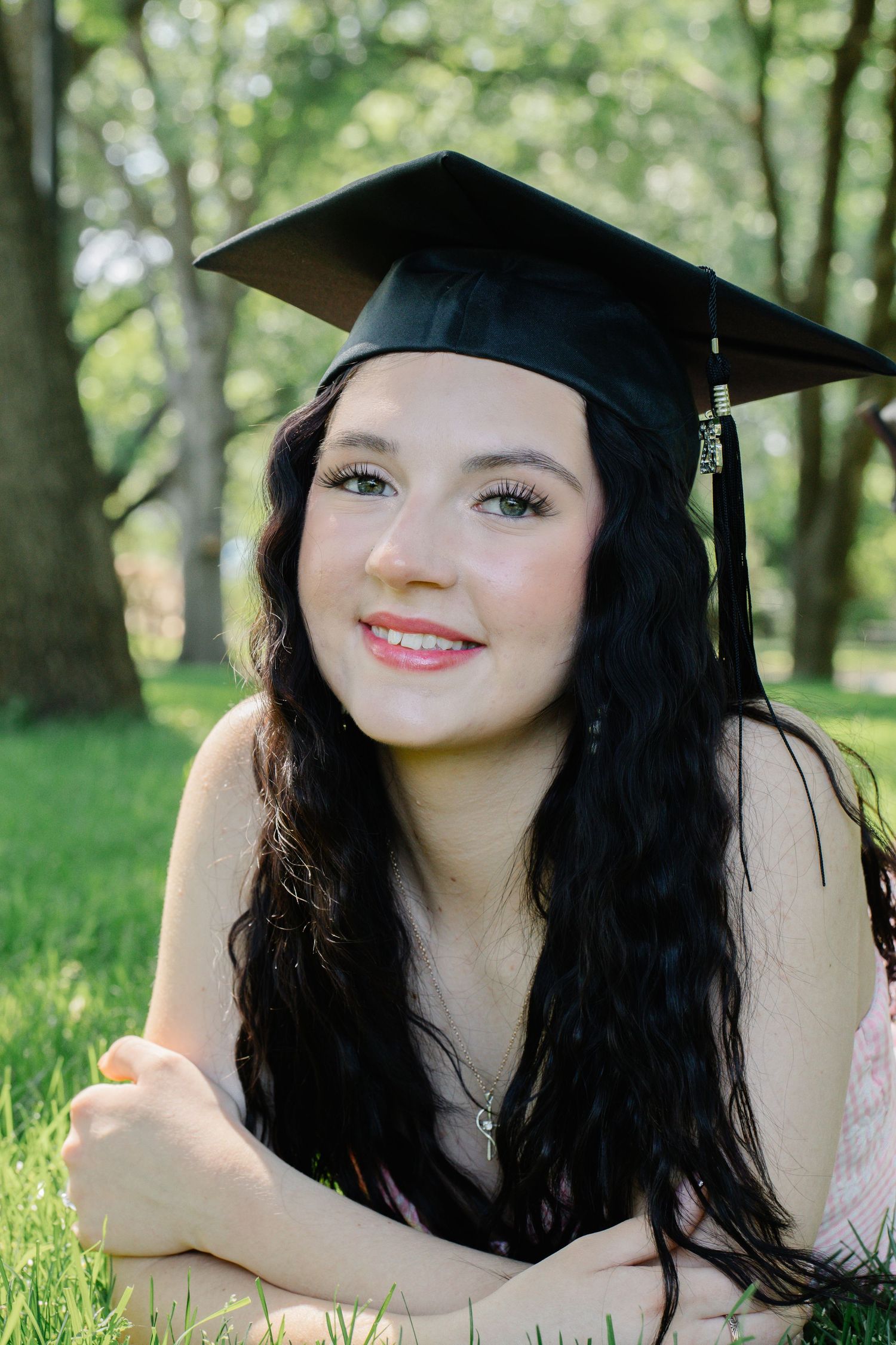 Smiling graduate with long dark hair wearing black cap, lying on grass among trees.
