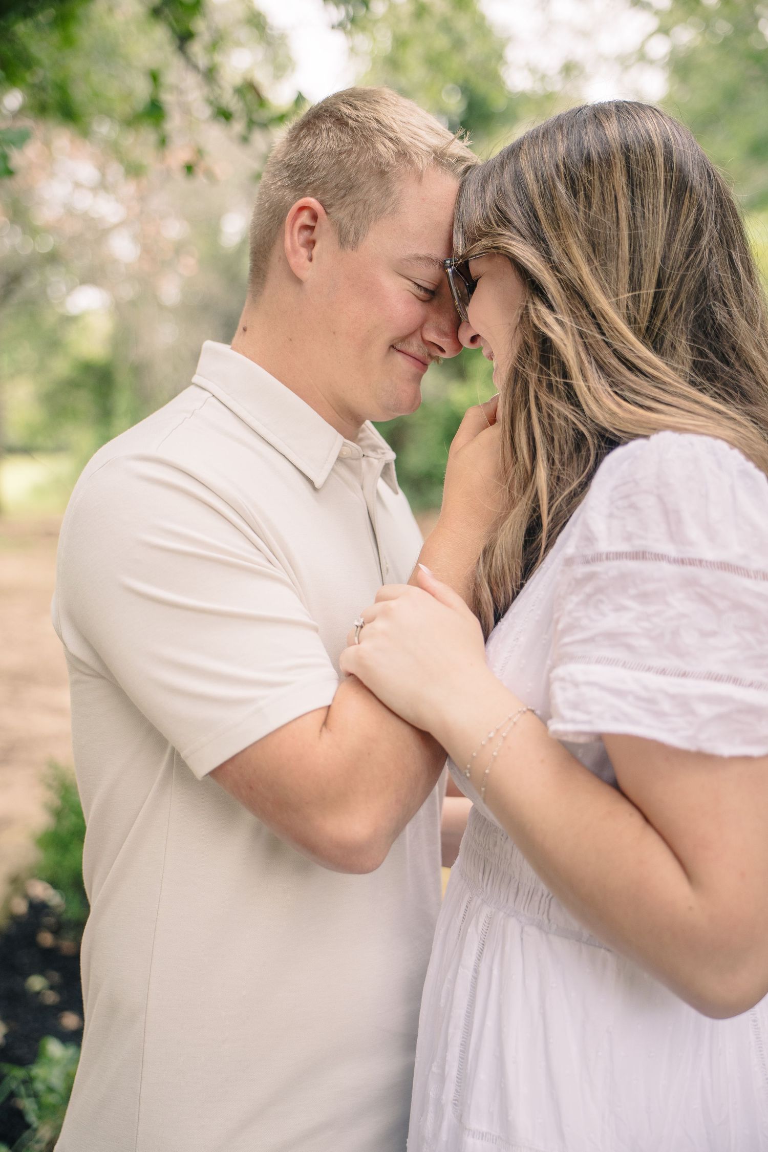 A couple shares a tender forehead-to-forehead moment outdoors, dressed in white and cream attire, surrounded by lush greenery.