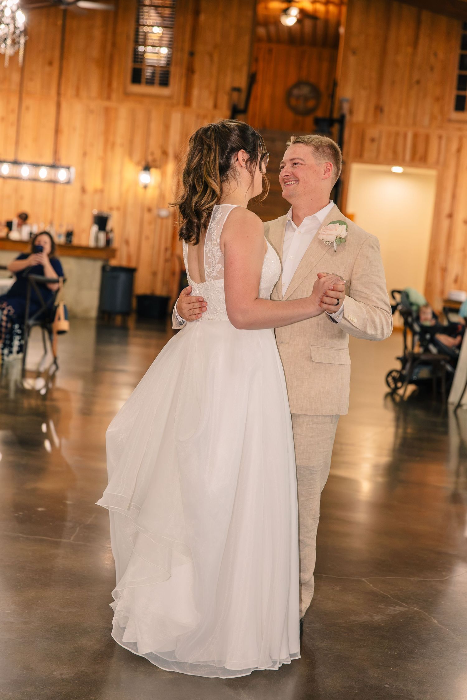 Bride in a white gown and groom in a tan suit share their first dance in a rustic wooden barn wedding venue.
