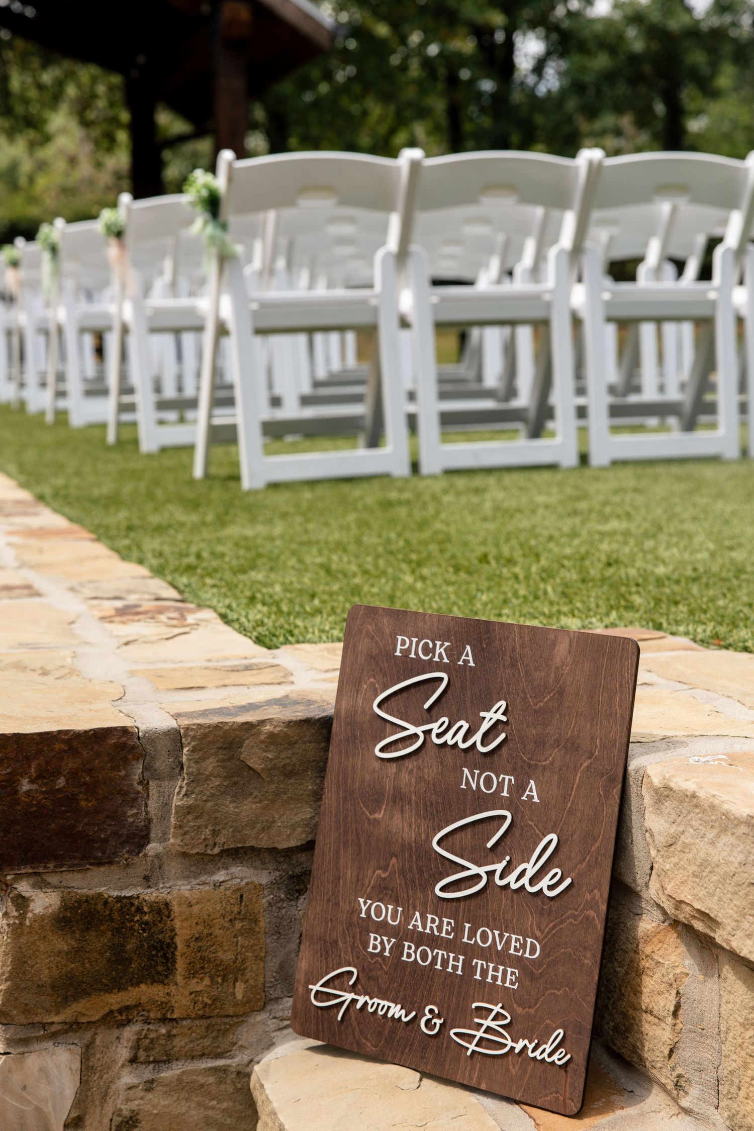Wooden wedding sign reading Pick a Seat Not a Side beside white folding chairs on a green lawn at an outdoor ceremony.