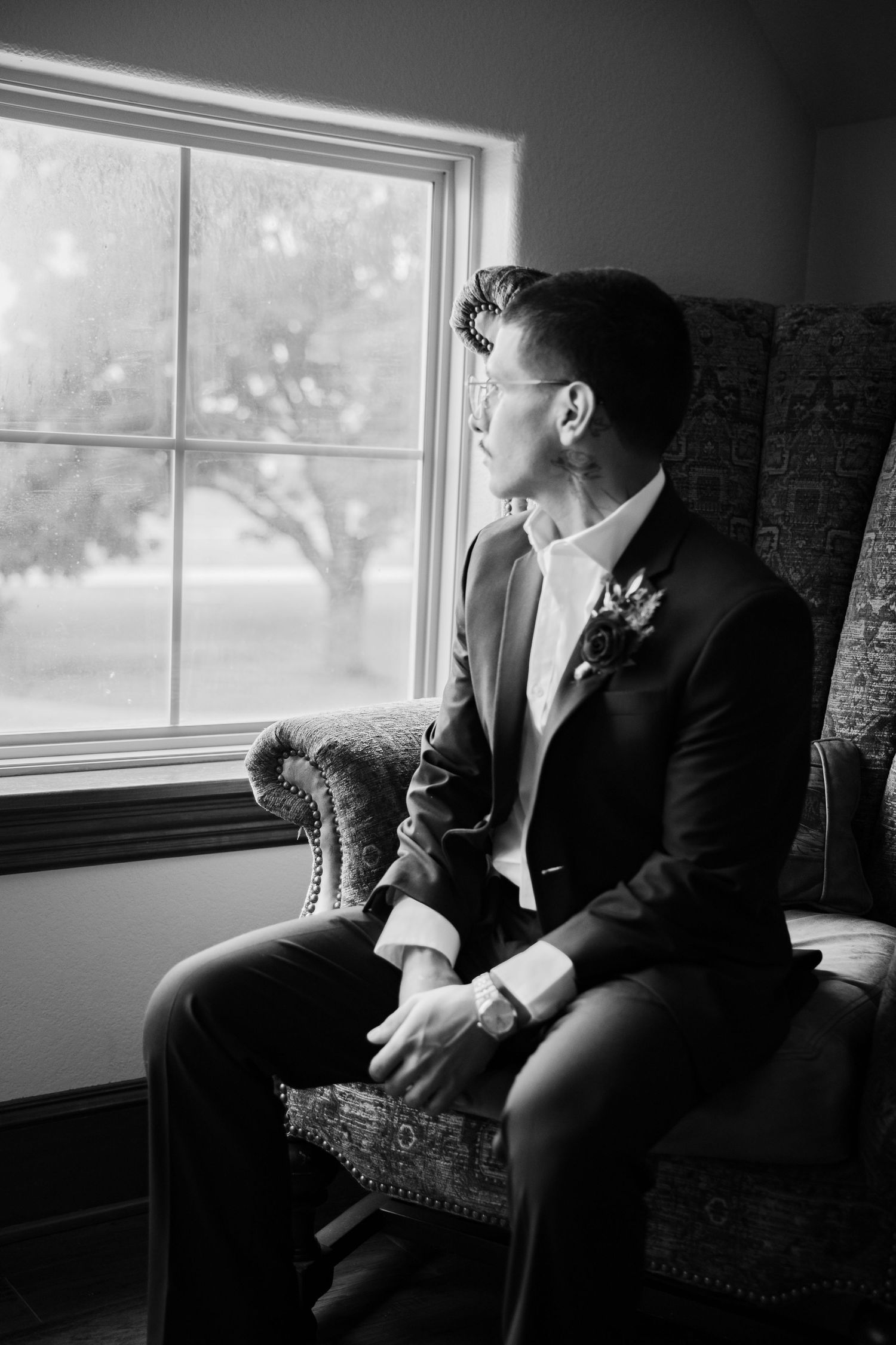 Black and white portrait of a groom in a suit with a sitting in a patterned armchair, gazing out a sunlit window. Burleson Tx